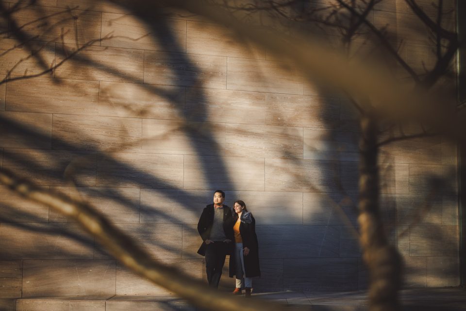 Two people stand against a sunlit stone wall, with tree branches and their shadows in the foreground. Two people stand against a sunlit stone wall, with tree branches and their shadows in the foreground.