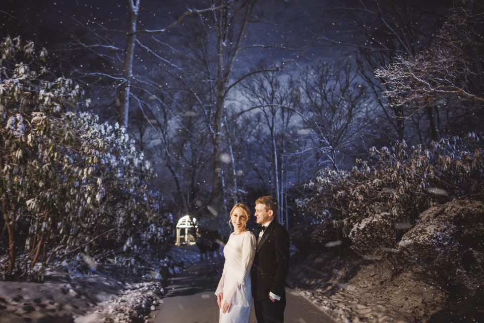 A bride and groom stand together on a snowy path at night, surrounded by snow-covered trees and falling snowflakes. A bride and groom stand together on a snowy path at night, surrounded by snow-covered trees and falling snowflakes.