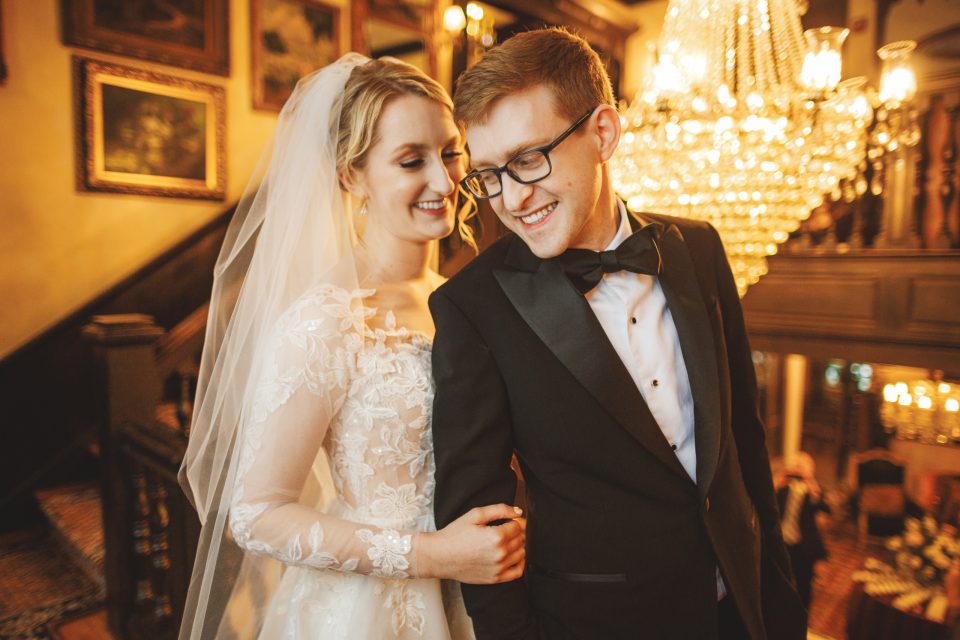 A bride in a lace wedding dress and veil stands arm in arm with a groom in a tuxedo, both smiling, in an ornate room with a chandelier and framed paintings. A bride in a lace wedding dress and veil stands arm in arm with a groom in a tuxedo, both smiling, in an ornate room with a chandelier and framed paintings.