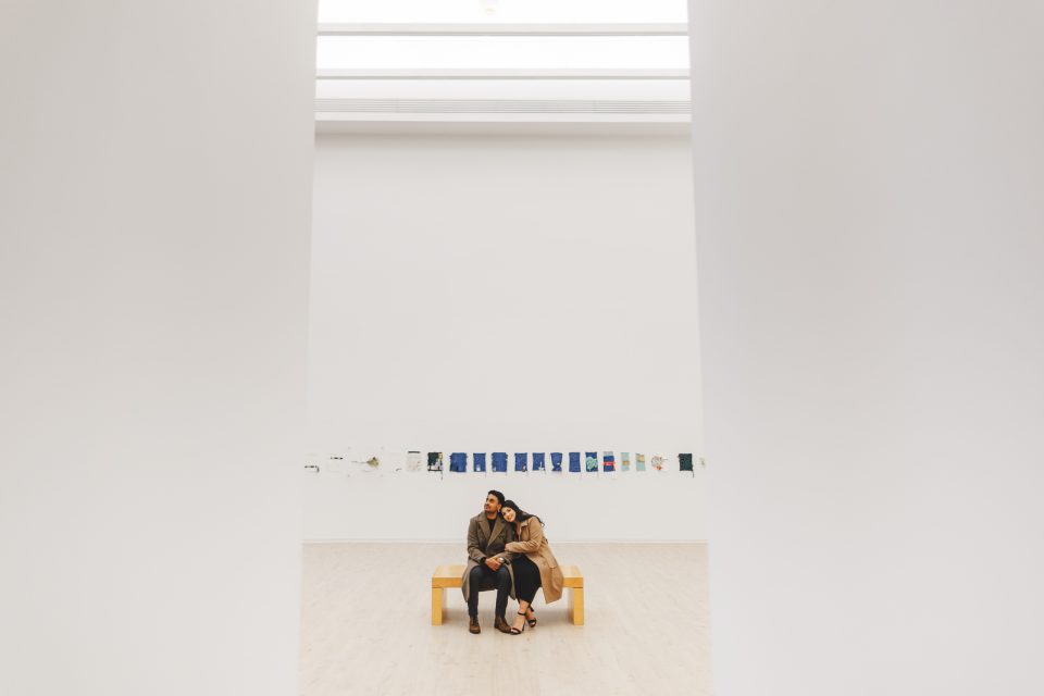 Two people sit closely together on a wooden bench in a spacious, minimalist art gallery with artworks displayed on the far white wall. Two people sit closely together on a wooden bench in a spacious, minimalist art gallery with artworks displayed on the far white wall.