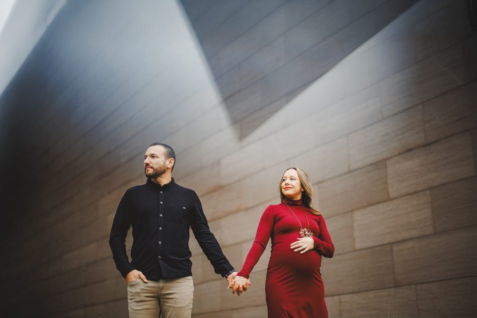 A man and a pregnant woman in a red dress hold hands while standing under angled light against a modern, gray stone wall. A man and a pregnant woman in a red dress hold hands while standing under angled light against a modern, gray stone wall.