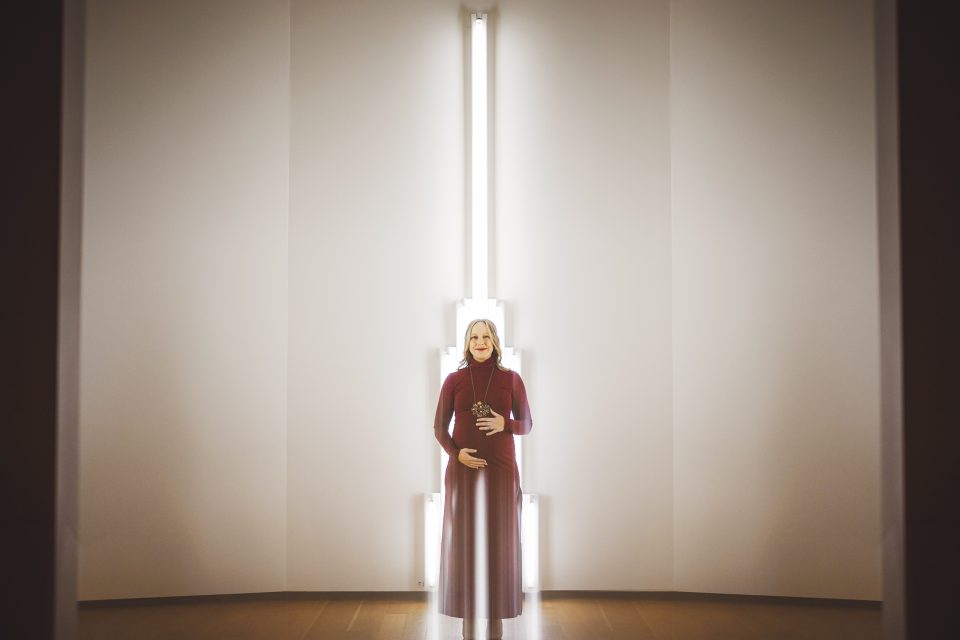 A person in a long red dress stands in front of a vertical light installation on a white wall, with hands resting on their midsection. A person in a long red dress stands in front of a vertical light installation on a white wall, with hands resting on their midsection.