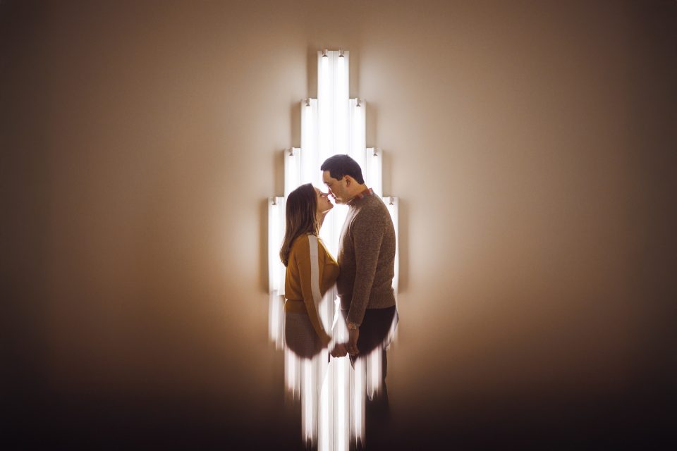 A couple stands close together in front of a wall-mounted vertical light fixture, holding hands and facing each other. The lights create a radiant, symmetrical backdrop. A couple stands close together in front of a wall-mounted vertical light fixture, holding hands and facing each other. The lights create a radiant, symmetrical backdrop.