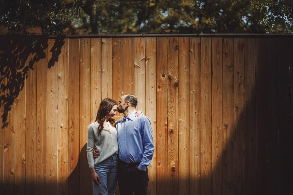 A couple stands close together in front of a wooden fence, with the man kissing the woman’s forehead. Sunlight casts a shadow on part of the scene. A couple stands close together in front of a wooden fence, with the man kissing the woman's forehead. Sunlight casts a shadow on part of the scene.