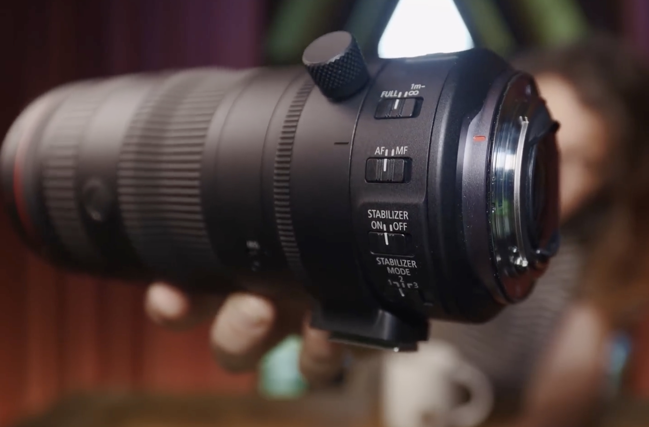 A close-up of a camera telephoto lens showing control switches for autofocus, stabilizer, and stabilizer mode, with a blurred background.