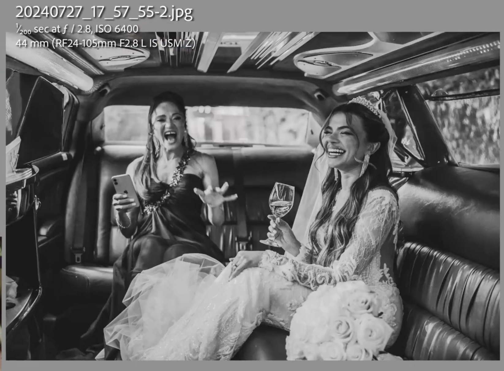 Two women in formal dresses sit in the back of a limousine, one holding a bouquet and wine glass, both smiling and laughing.