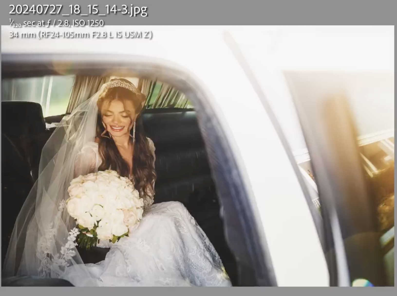 A bride in a white dress and veil, holding a bouquet of white flowers, sits in the back seat of a car, smiling and looking down.