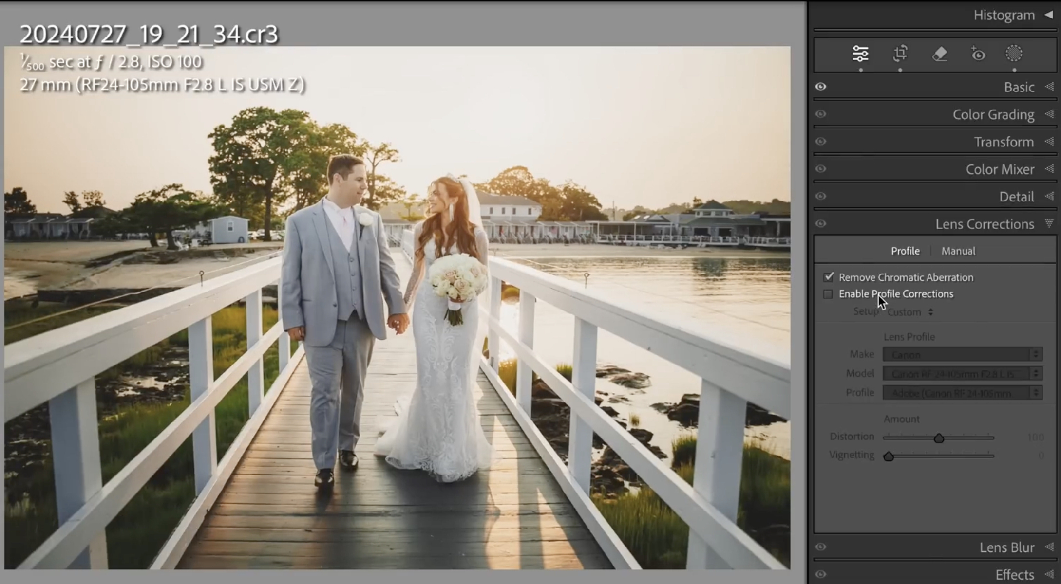 A bride and groom walk hand in hand on a wooden dock by the water at sunset, dressed in wedding attire, with houses and trees in the background.