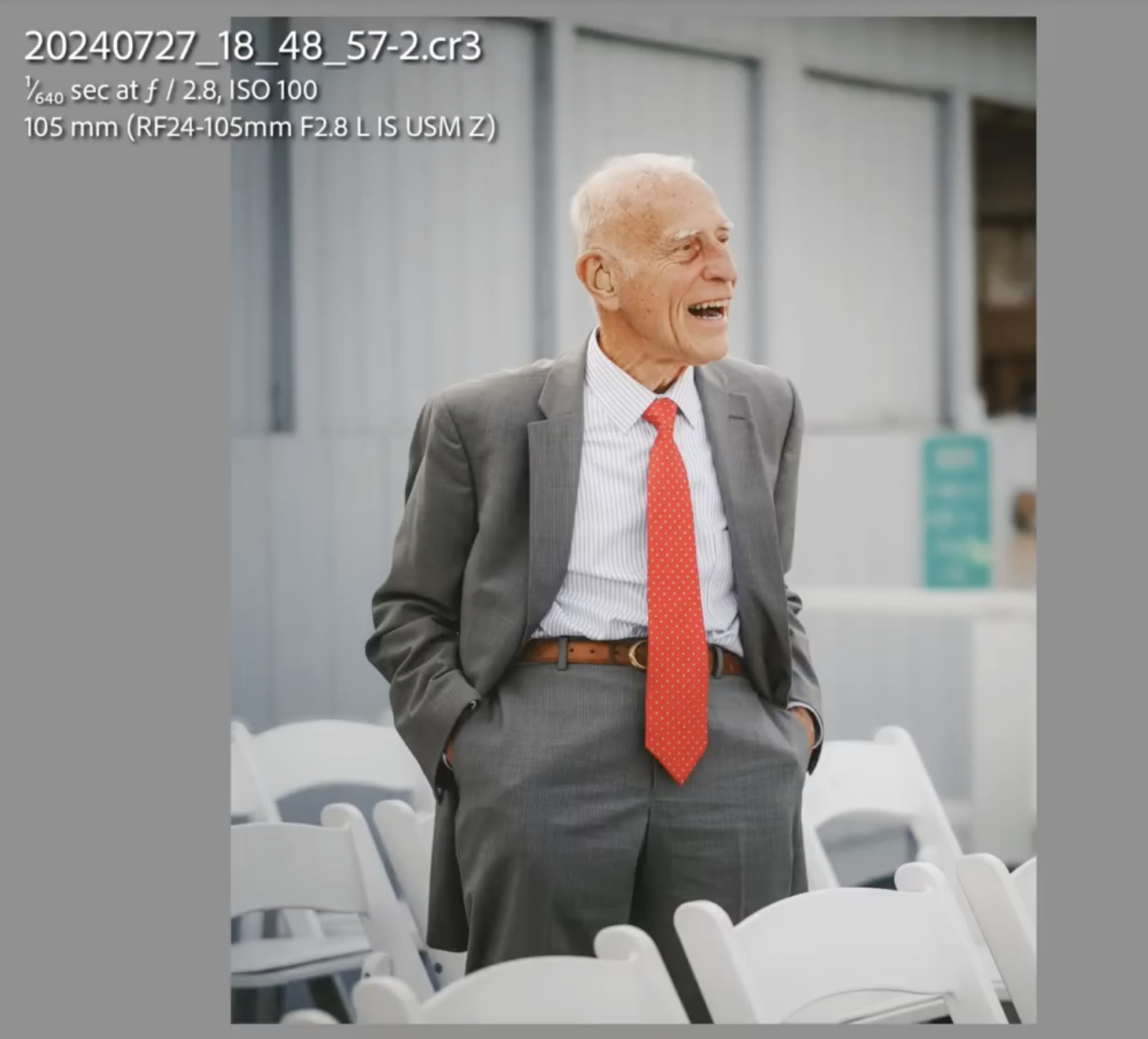An elderly man in a gray suit and red tie stands among white chairs outdoors, hands in pockets, smiling and looking to the side.