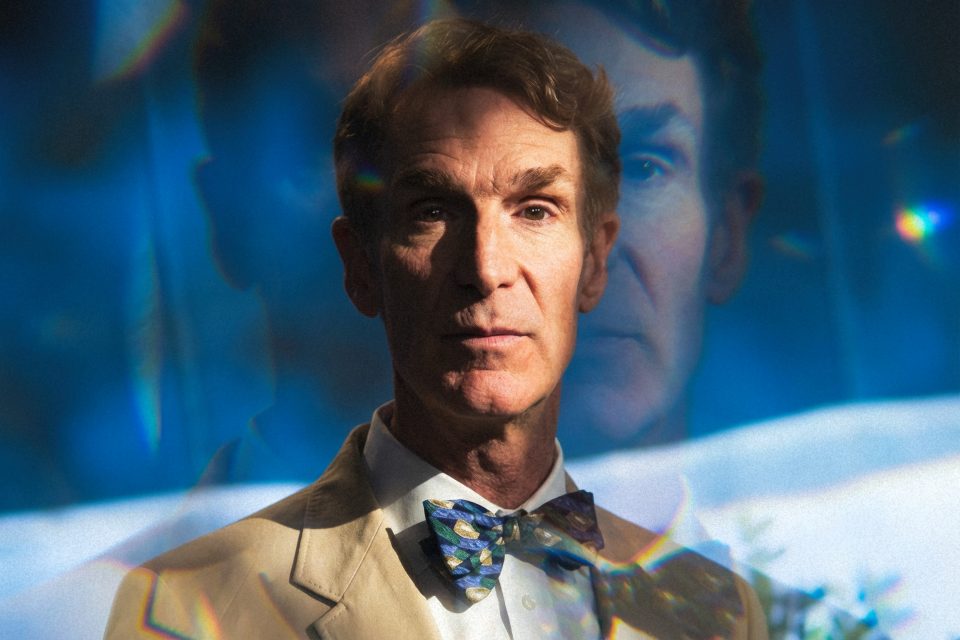 Bill Nye in a tan suit and patterned bow tie stands in front of a blue background with a double exposure effect showing his face twice, captured in striking detail by a dc portrait photographer. Bill Nye in a tan suit and patterned bow tie stands in front of a blue background with a double exposure effect showing his face twice, captured in striking detail by a dc portrait photographer.