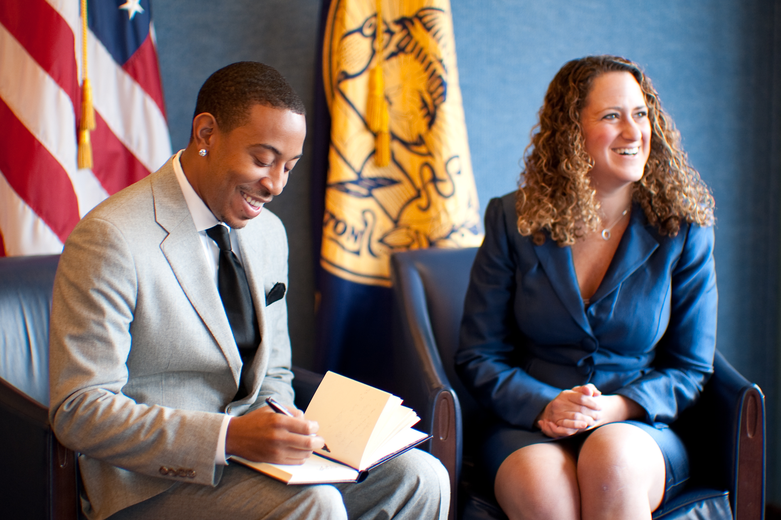 A man in a grey suit signs a notebook while sitting next to a woman in a blue suit; U.S. flags are visible in the background.