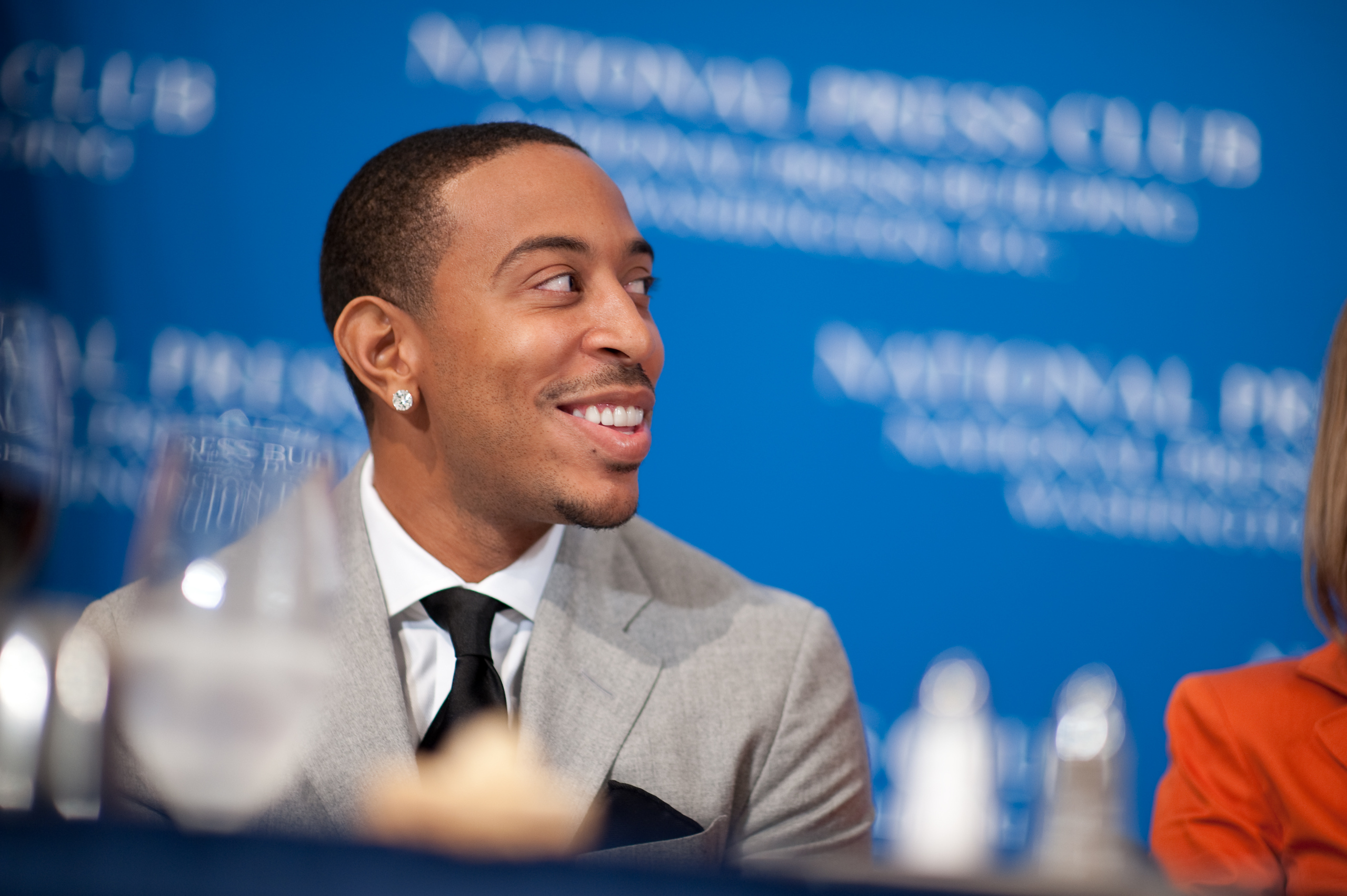 A man in a suit and tie smiles while seated at a table with a blue National Press Club backdrop behind him.