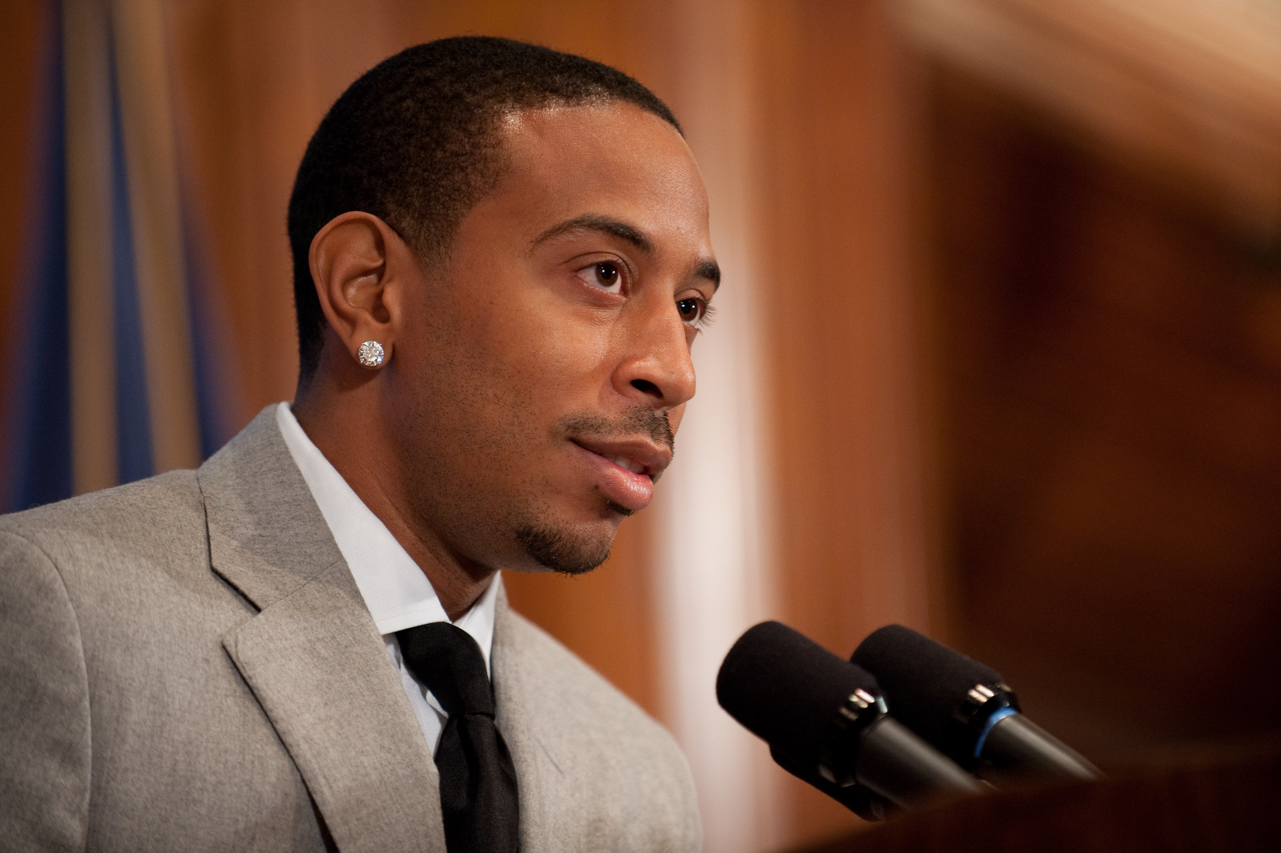 A man in a gray suit and black tie speaks into a microphone at a podium, with a blurred background behind him.