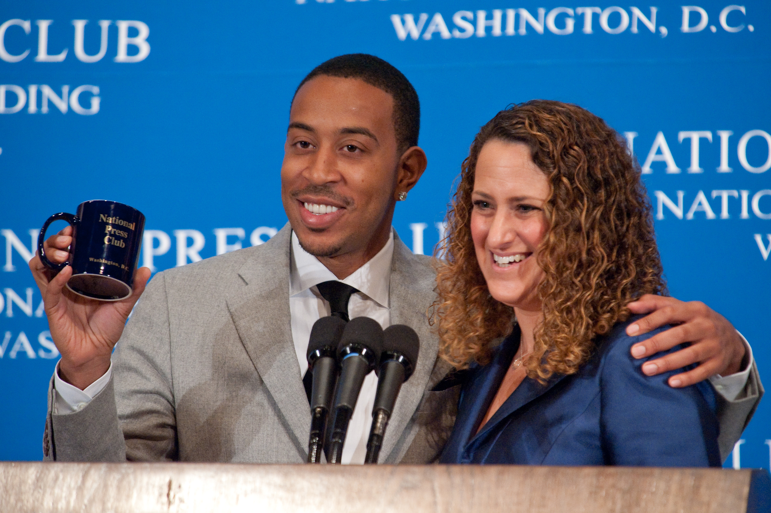 A man in a suit holds a National Press Club mug and stands with a smiling woman at a podium with microphones in front of a blue backdrop reading "National Press Club, Washington, D.C.