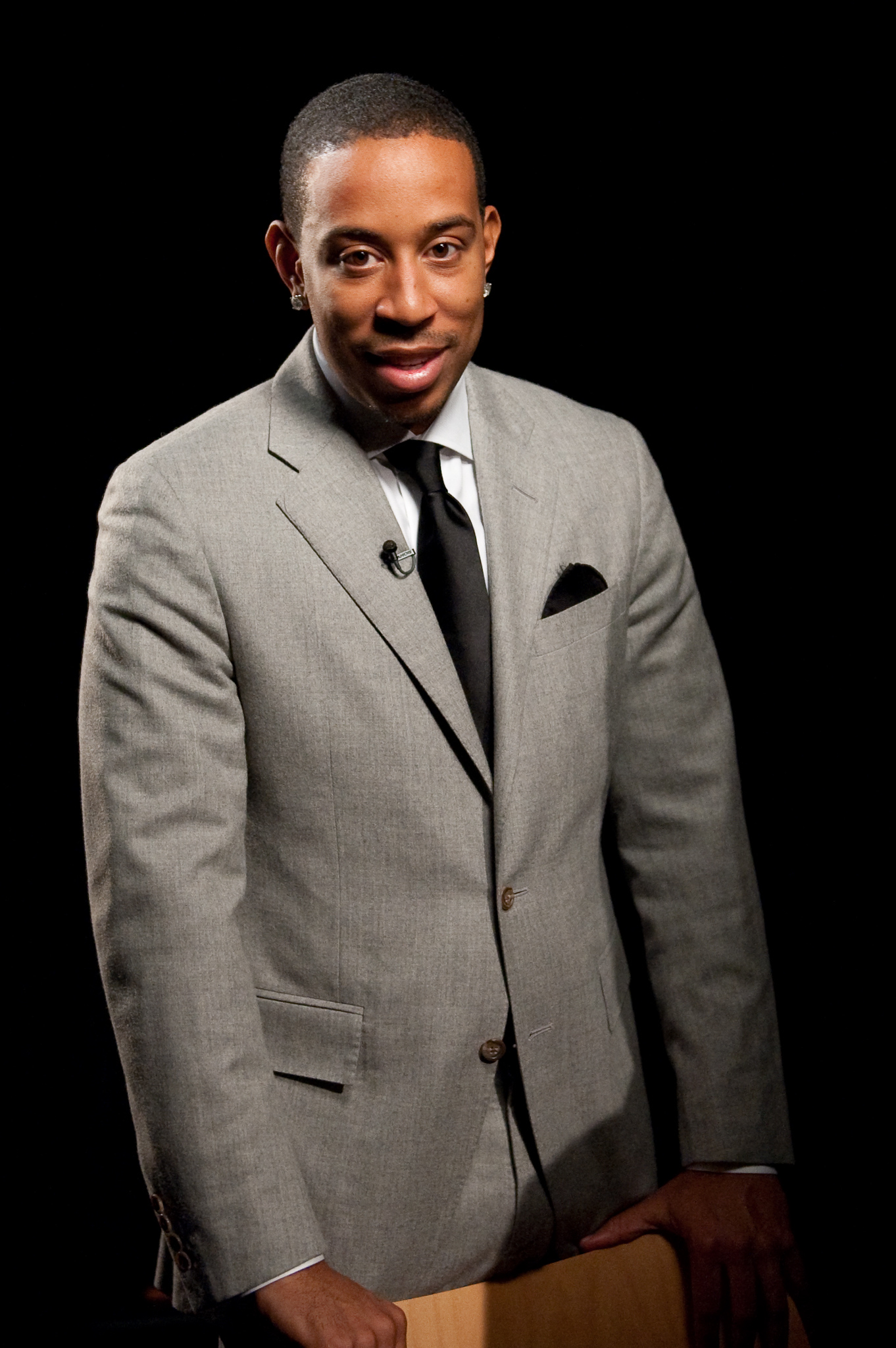 A man in a light gray suit and black tie stands and smiles at the camera against a dark background.