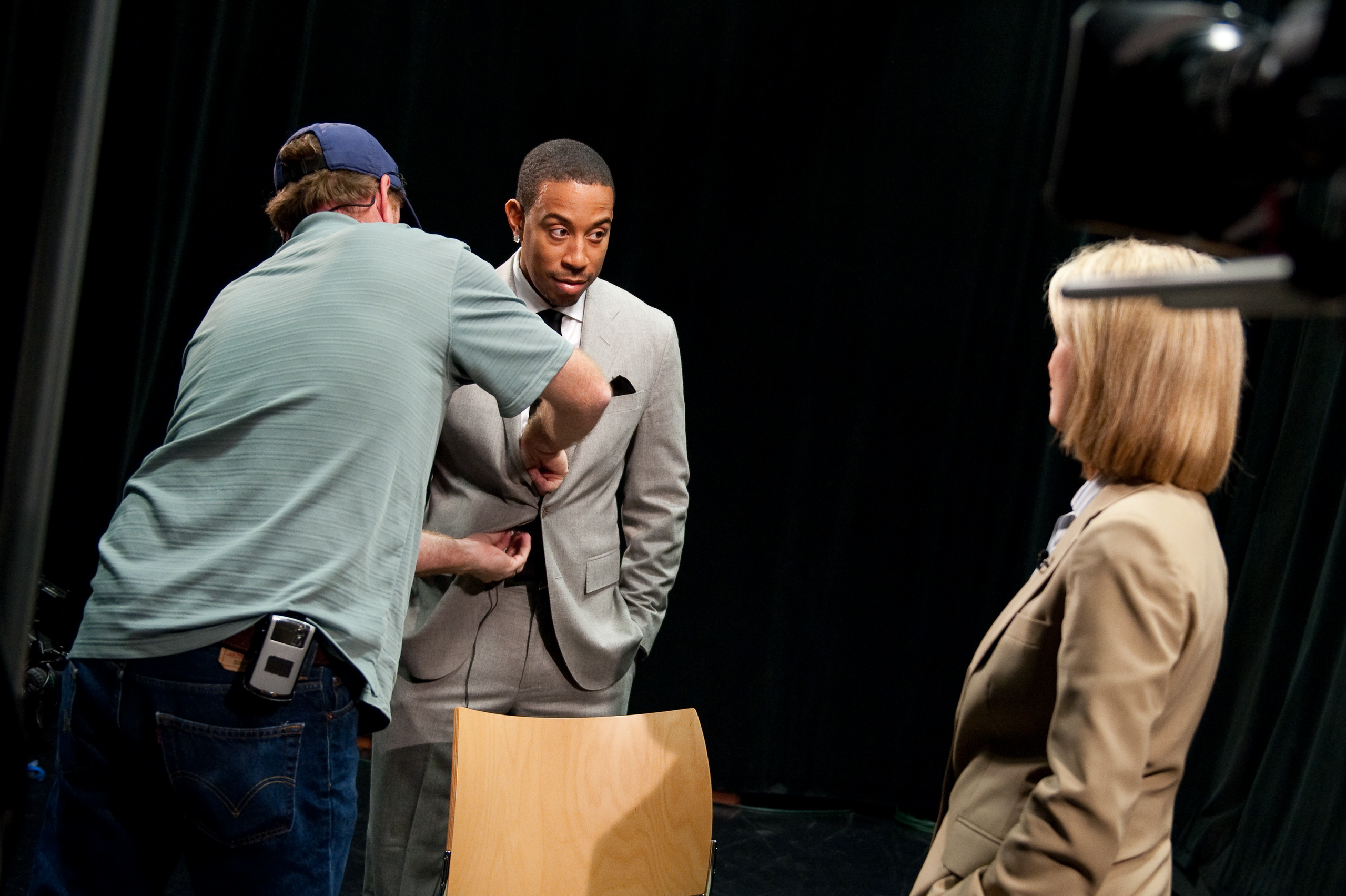 A man in a gray suit is being fitted with a microphone by a crew member while a woman in a tan blazer stands nearby in a studio setting.