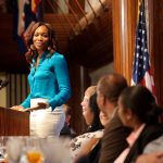 A woman stands at a podium speaking to an audience seated at a table, with flags and a National Press Club sign in the background.