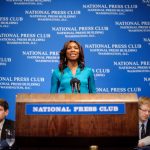 A woman stands at a podium labeled "National Press Club," speaking, with two men seated on either side, against a backdrop with the same text.