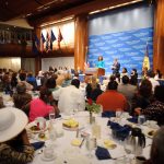 A large audience sits at round tables in a banquet hall, listening to speakers on a stage with a blue backdrop and national flags.