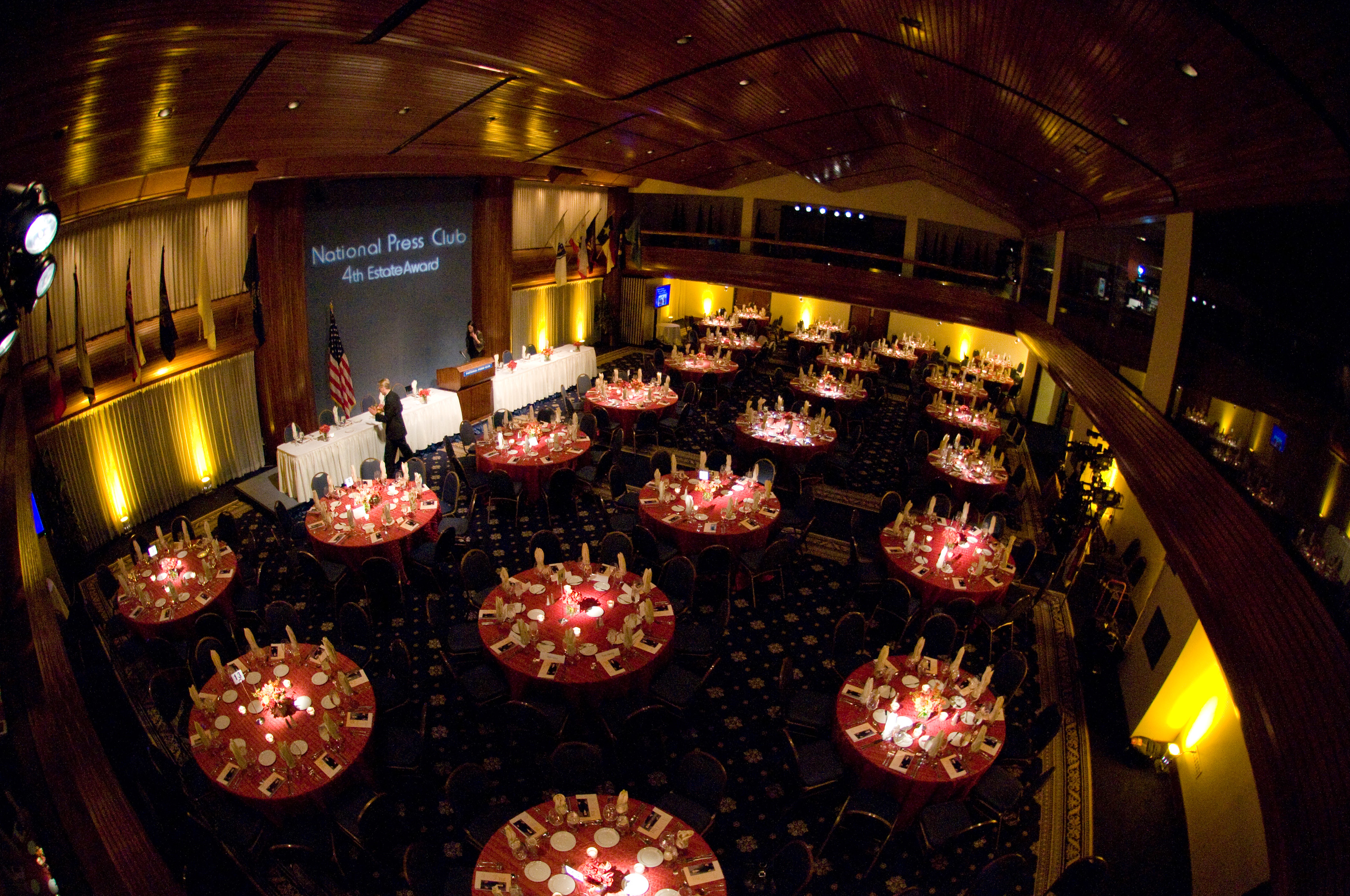 Banquet hall with round tables set for an event at the National Press Club, featuring a stage, American flag, and dim, warm lighting.