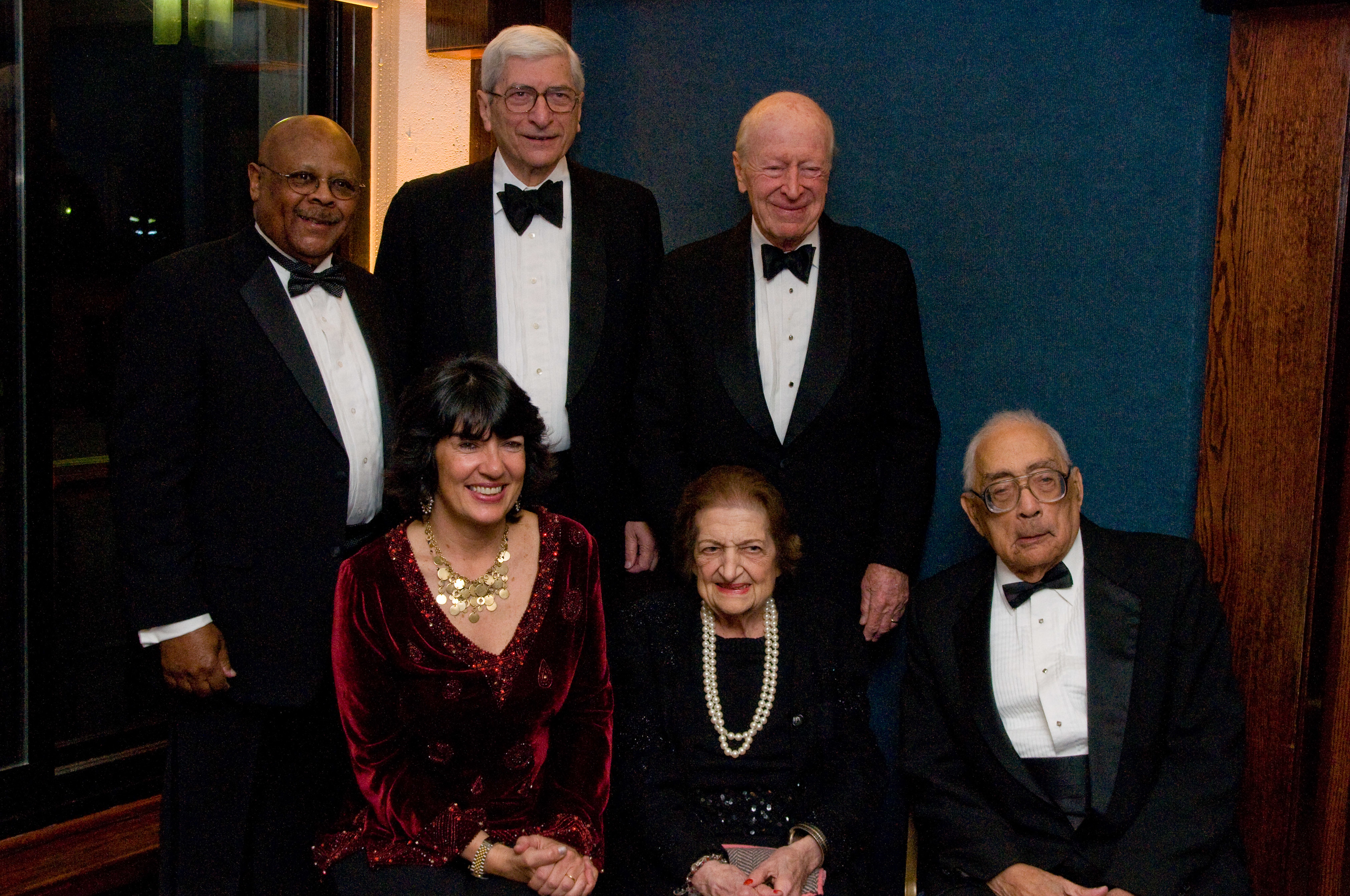 Six adults, four men and two women, dressed in formal evening wear pose together indoors against a blue and brown background.