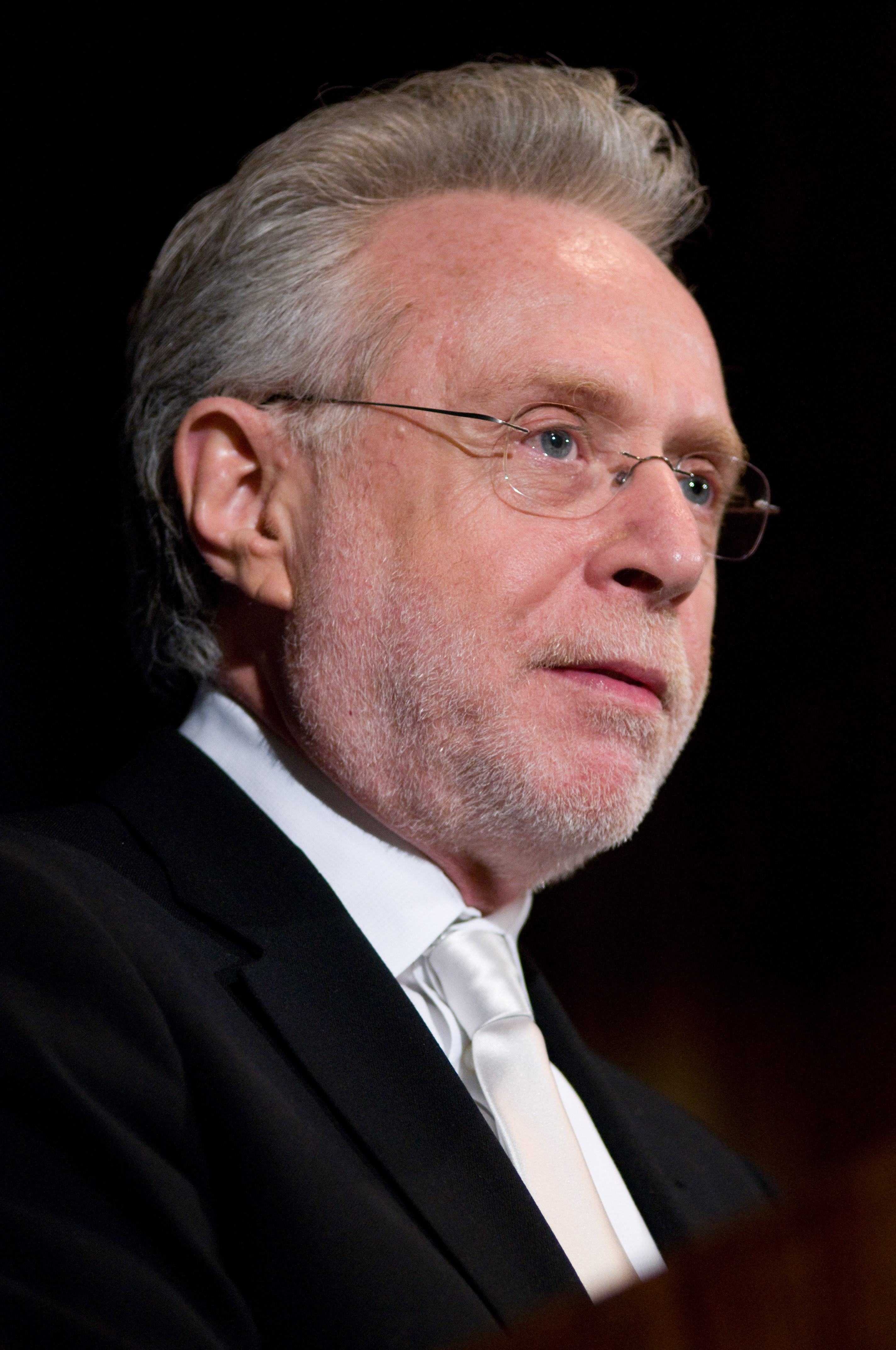 A man with gray hair, glasses, and a beard is wearing a black suit with a white shirt and tie, looking slightly to the side.