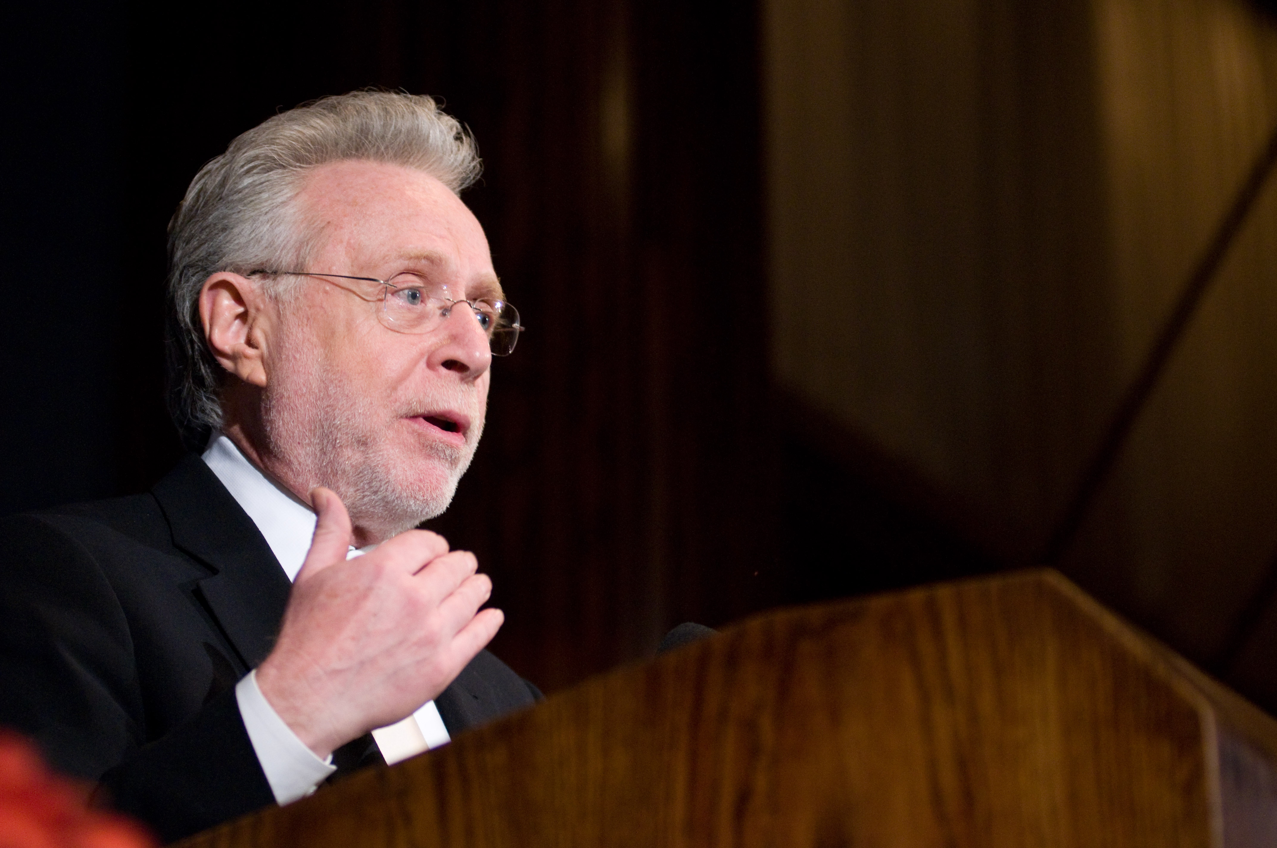 A man in a suit and glasses speaks at a podium, gesturing with his left hand.