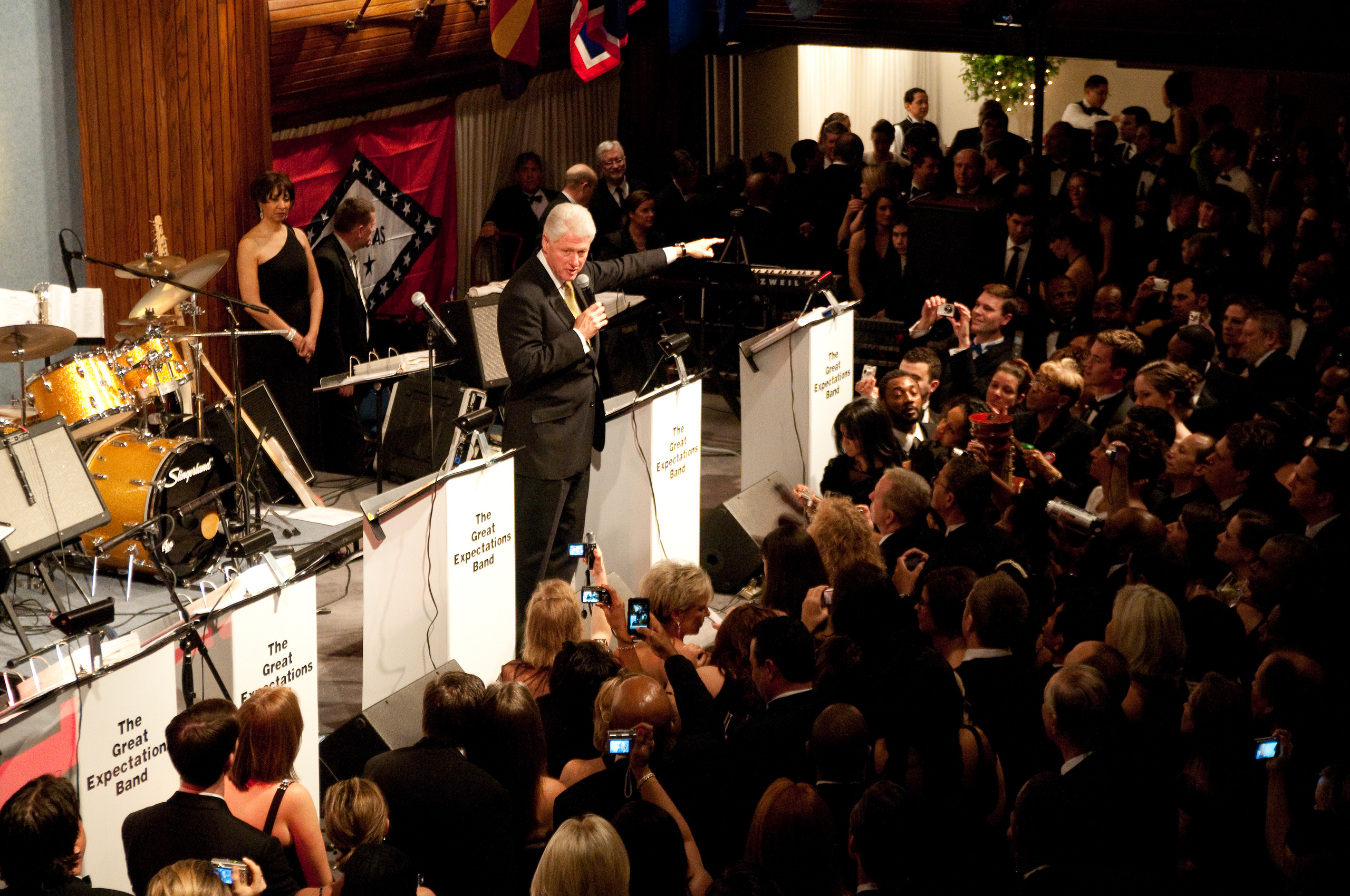 A man in a suit speaks on stage with a microphone at a crowded indoor event; a band setup and several people with cameras are visible.