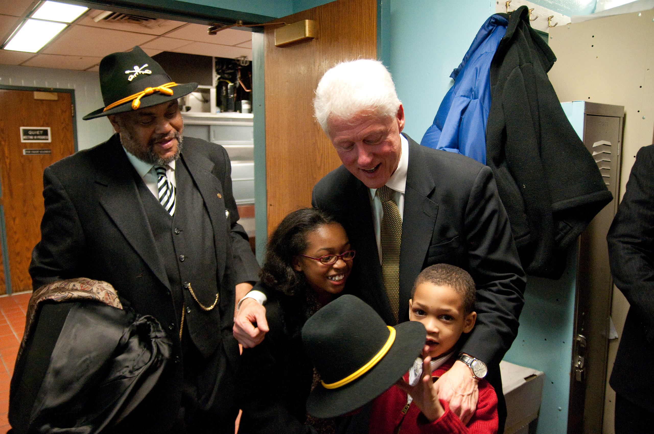 An adult man in a suit stands with two children and another man in a suit and hat in a hallway, with coats hanging on hooks in the background.