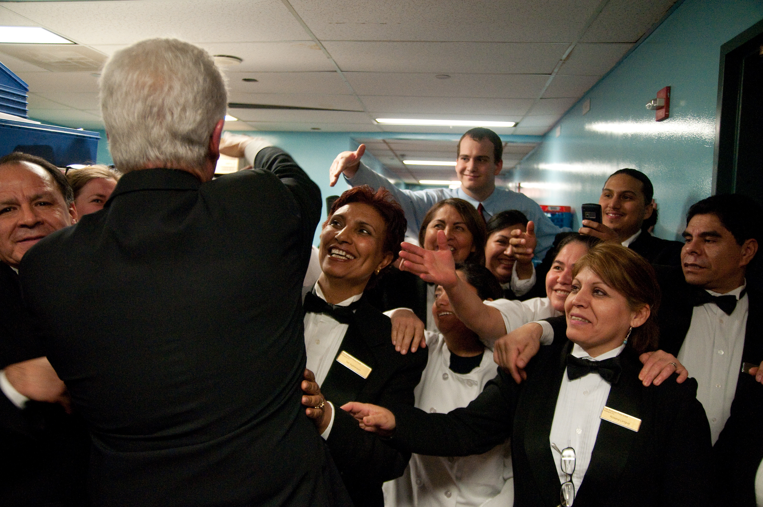 A group of formally dressed staff members smile and reach out as a man with gray hair stands in front of them in a hallway.