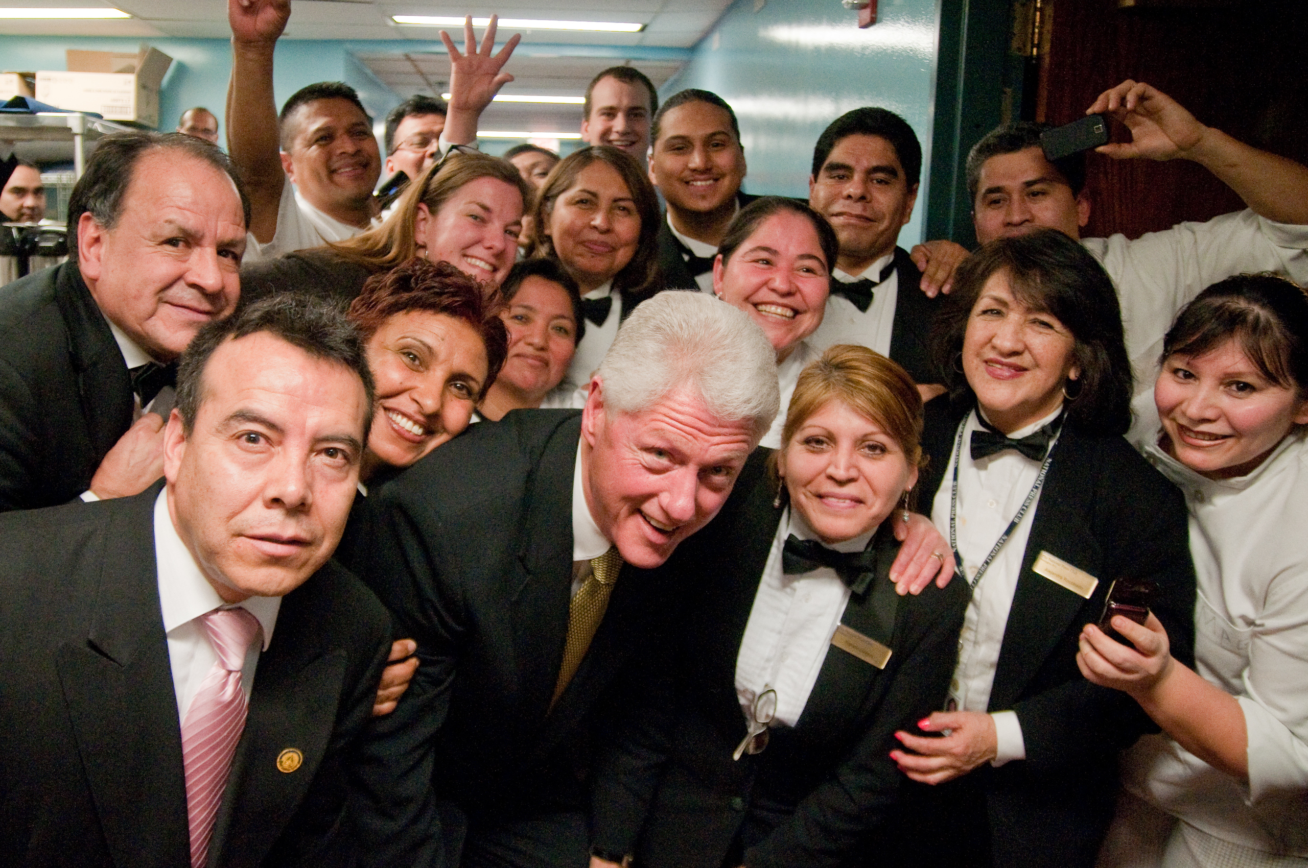 A group of restaurant staff in formal uniforms pose for a cheerful group photo with a man in a suit in the center.