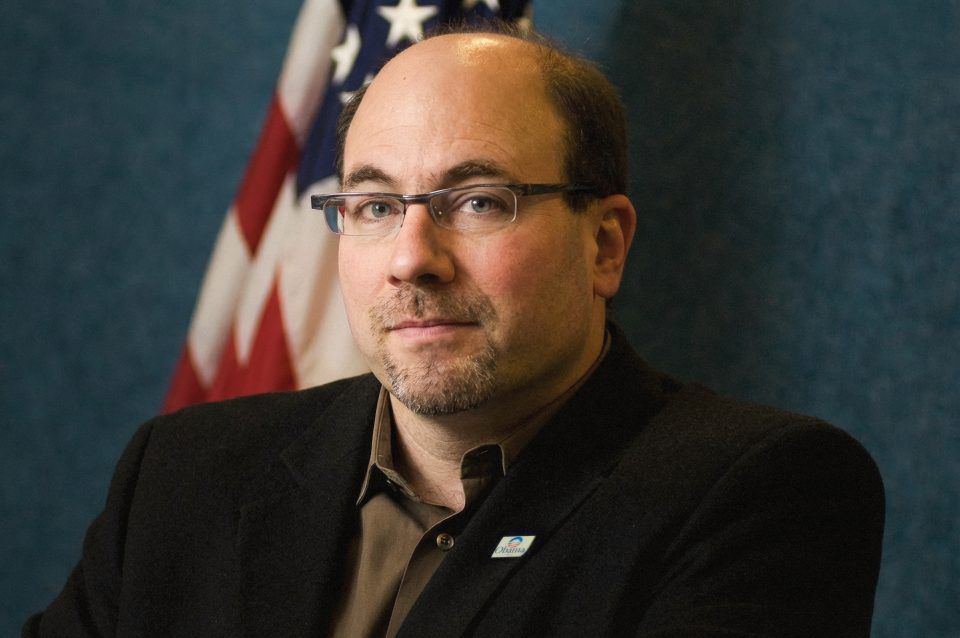 Craig Newmark, in this epic portrait, sits in front of a U.S. flag and a blue background, wearing glasses, a dark blazer, and a brown shirt. Craig Newmark, in this epic portrait, sits in front of a U.S. flag and a blue background, wearing glasses, a dark blazer, and a brown shirt.