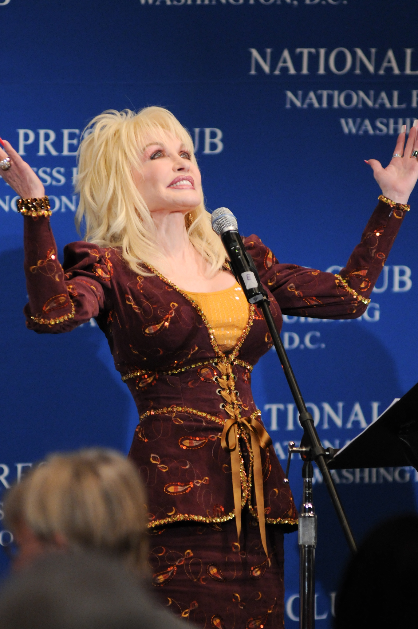 A woman with blonde hair in a brown and gold outfit stands at a podium with a microphone, raising her arms, in front of a blue backdrop reading "National Press Club, Washington D.C.