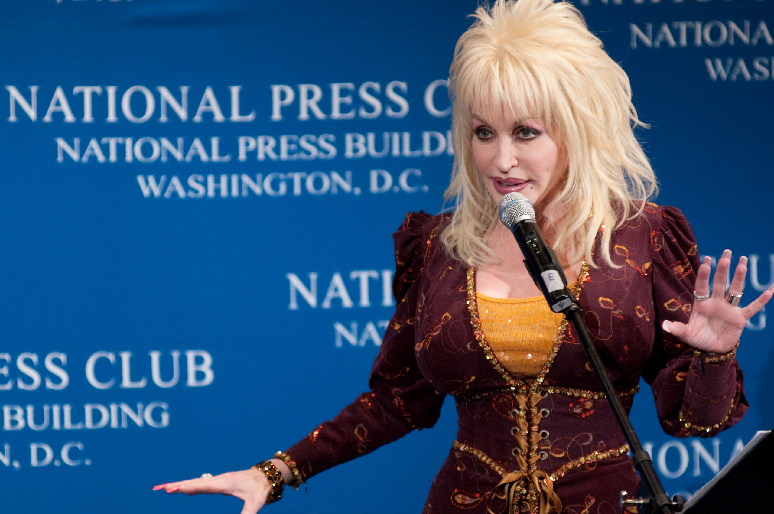A woman with blonde hair speaks at a podium with a microphone, standing in front of a blue National Press Club backdrop in Washington, D.C.