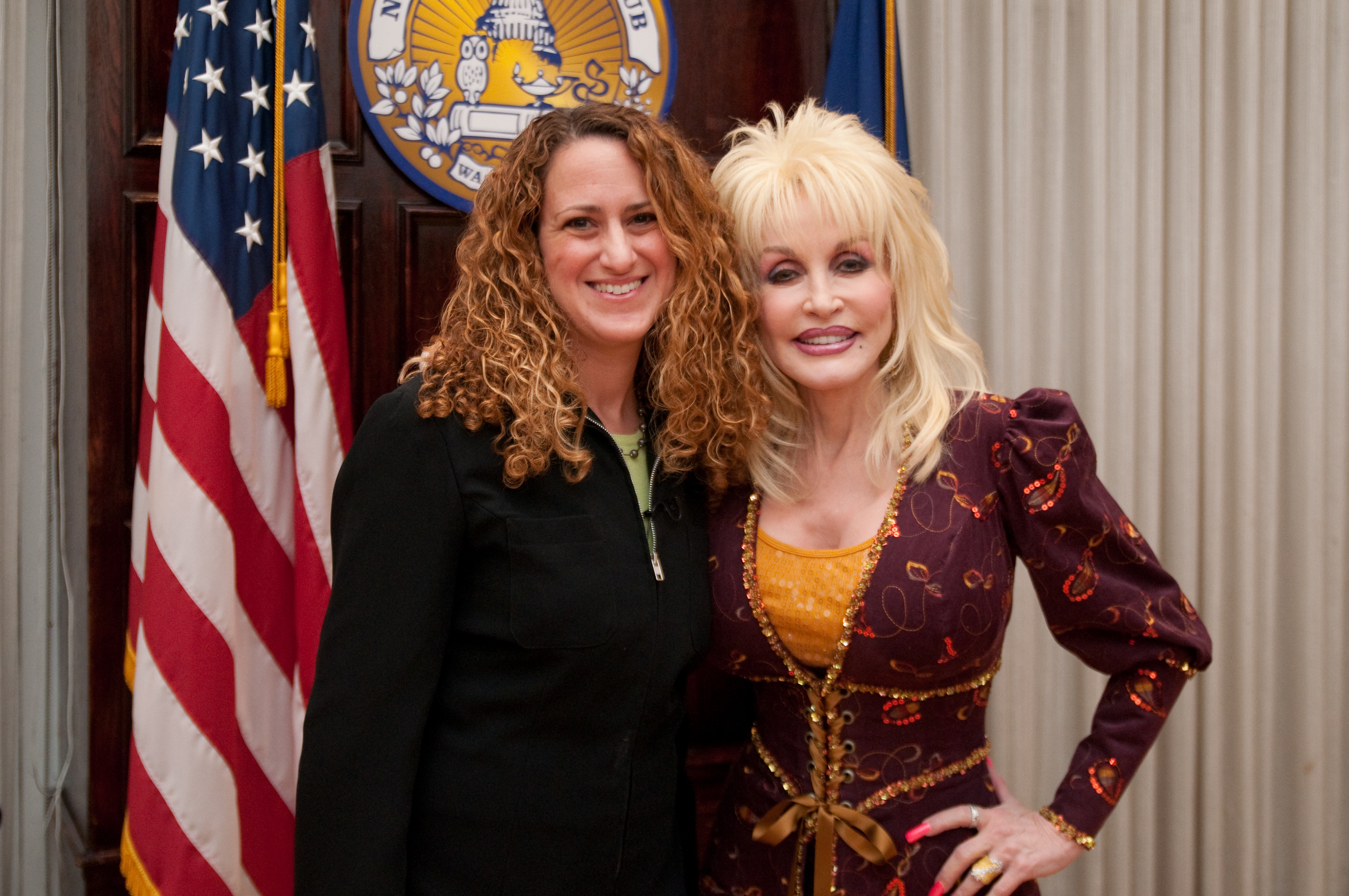 Two women stand side by side smiling in front of U.S. and state flags, with one woman wearing a patterned dress and the other in a black jacket.
