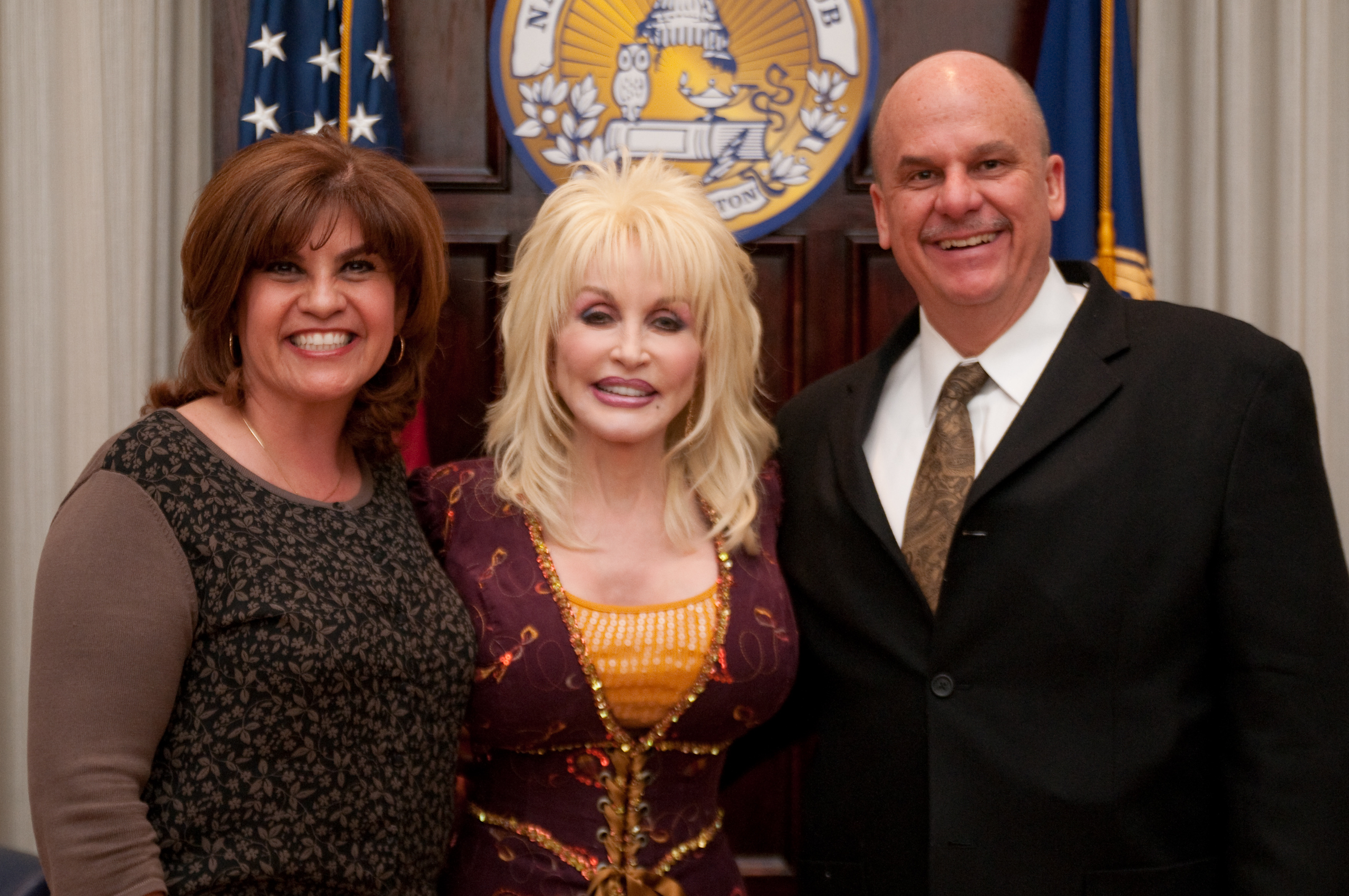 Three adults stand smiling in front of a wooden panel with an official seal and flags visible in the background.