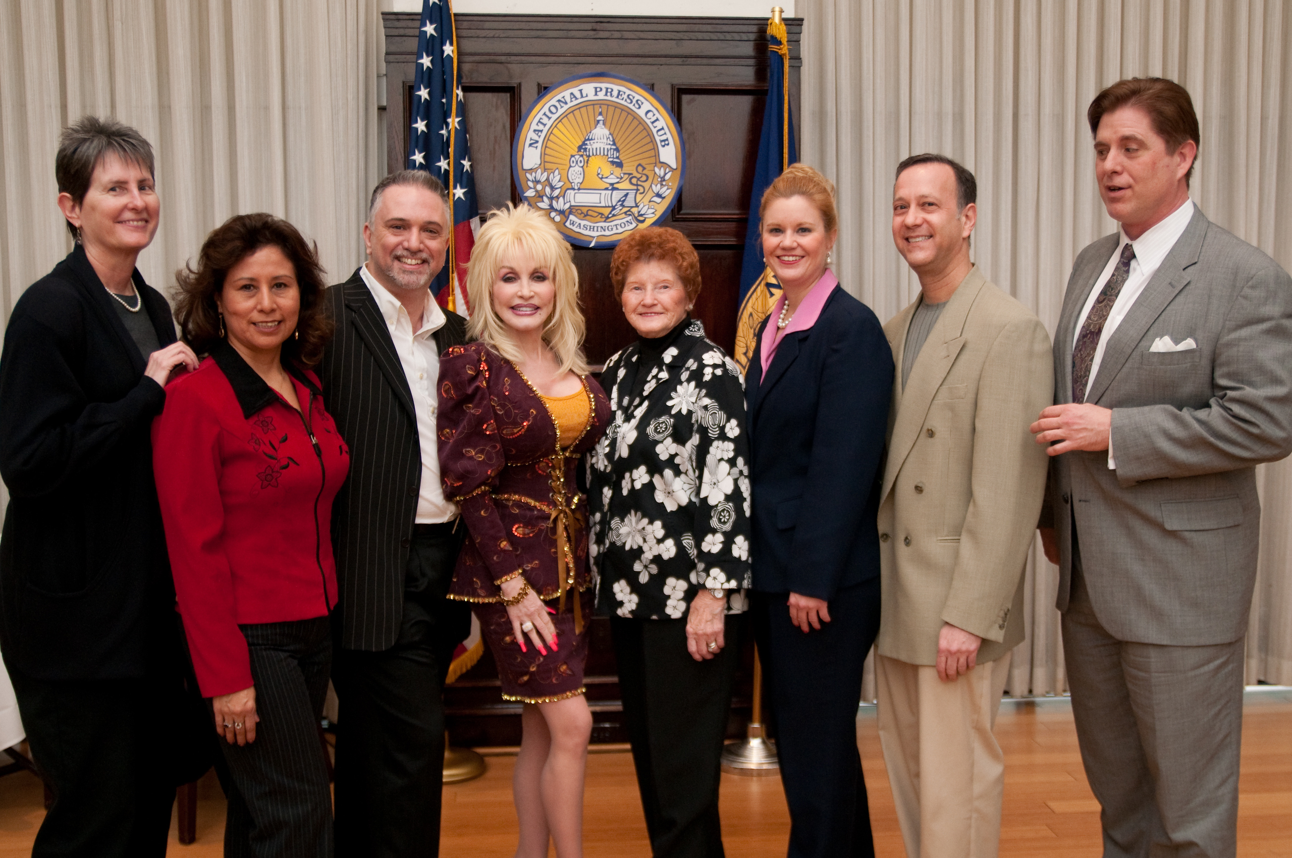 A group of seven adults pose together in front of an American flag and a sign reading “National Press Club,” smiling for the camera.