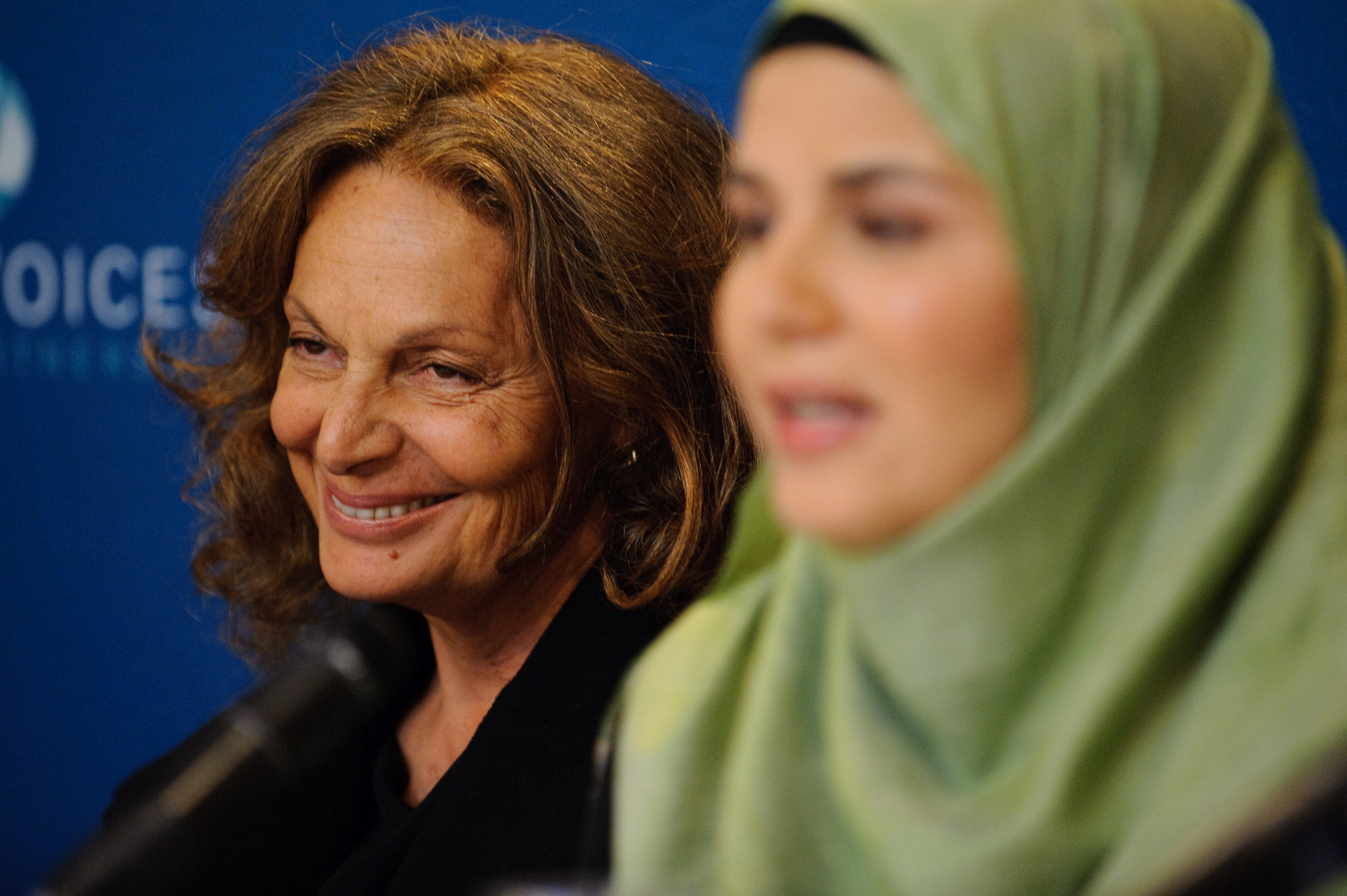 Two women sit side by side at a press conference; one is smiling and the other, wearing a green hijab, speaks into a microphone. A blue backdrop is behind them.