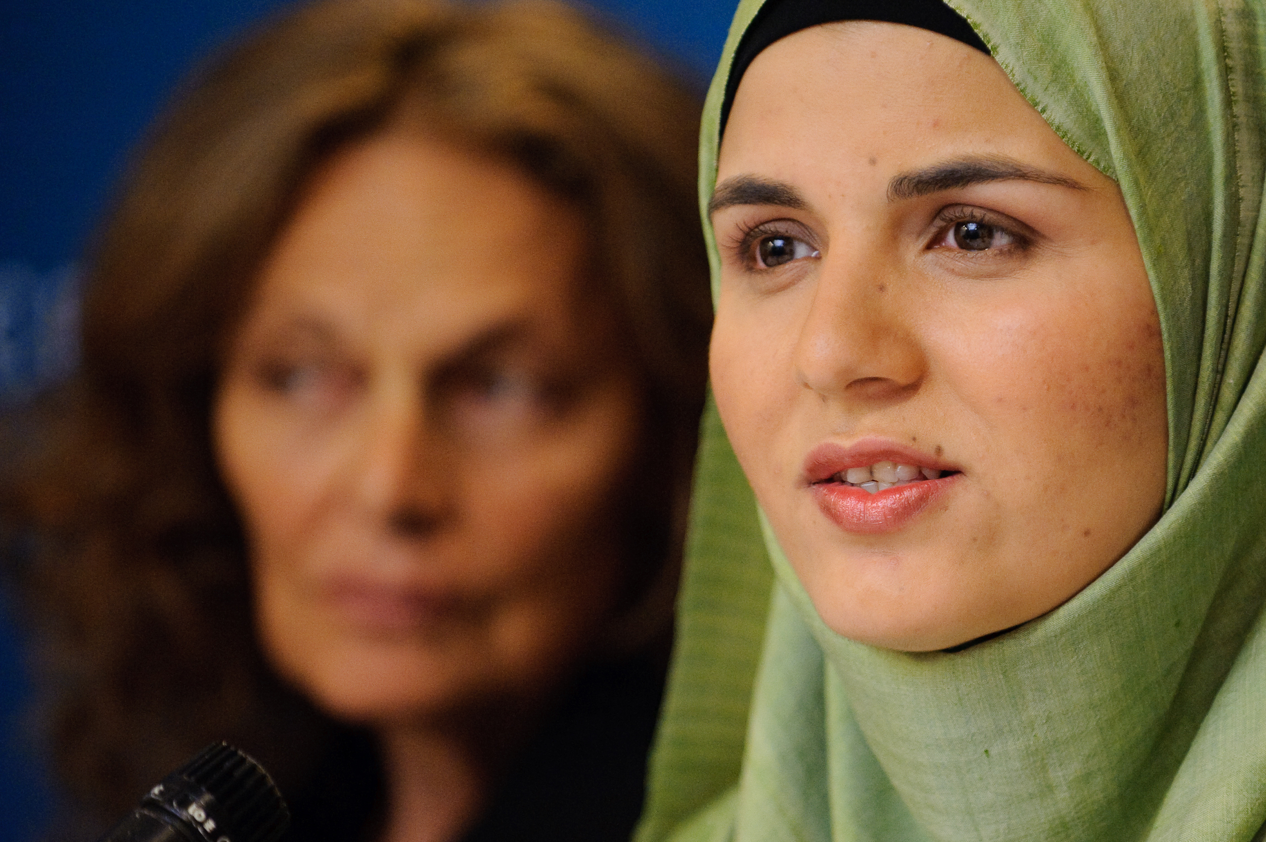 A woman in a green hijab speaks into a microphone while another woman in the background listens attentively.