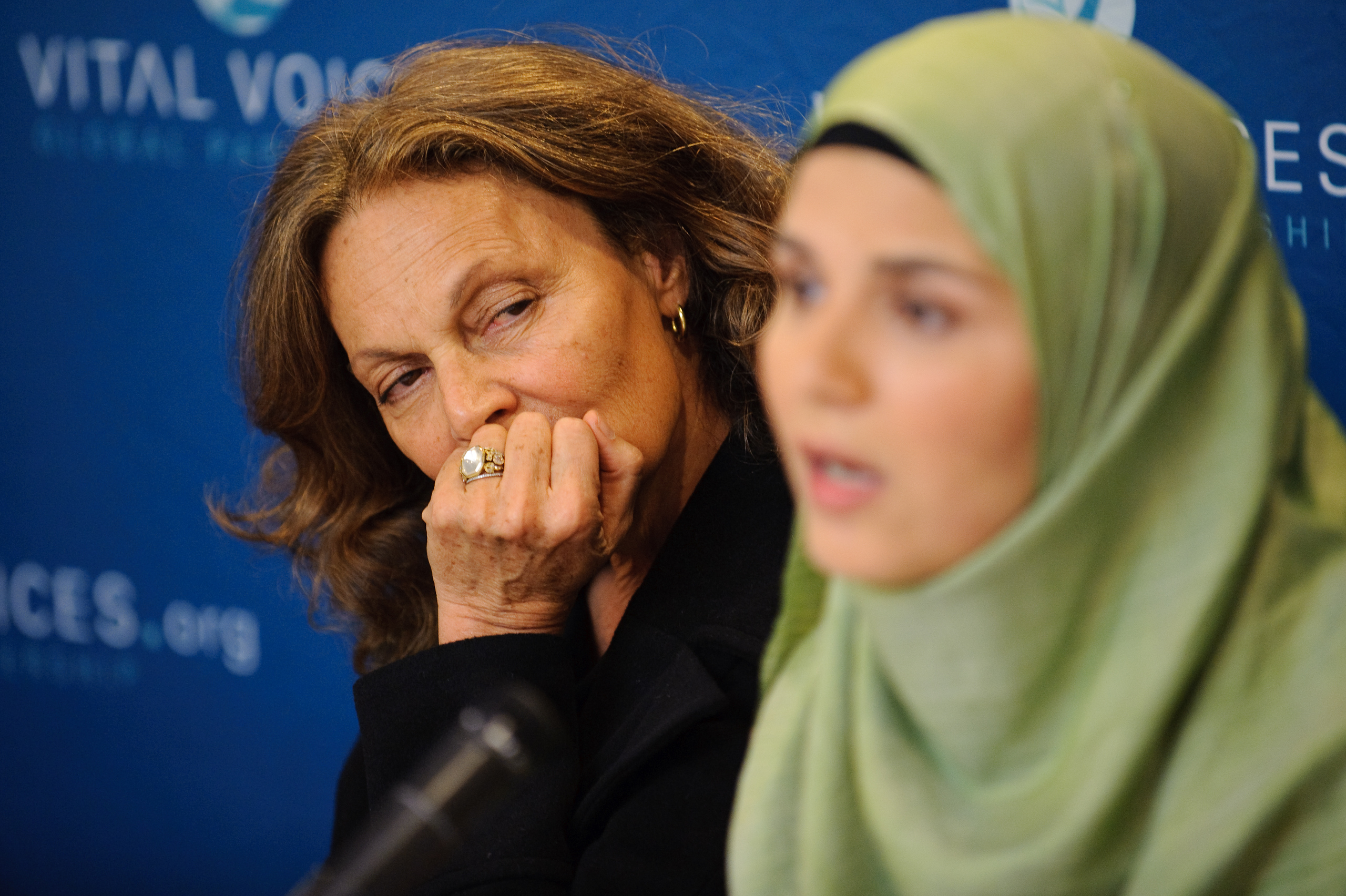 Two women sit in front of a blue "Vital Voices" backdrop; one woman in focus listens intently while the other, in a green hijab, speaks into a microphone.