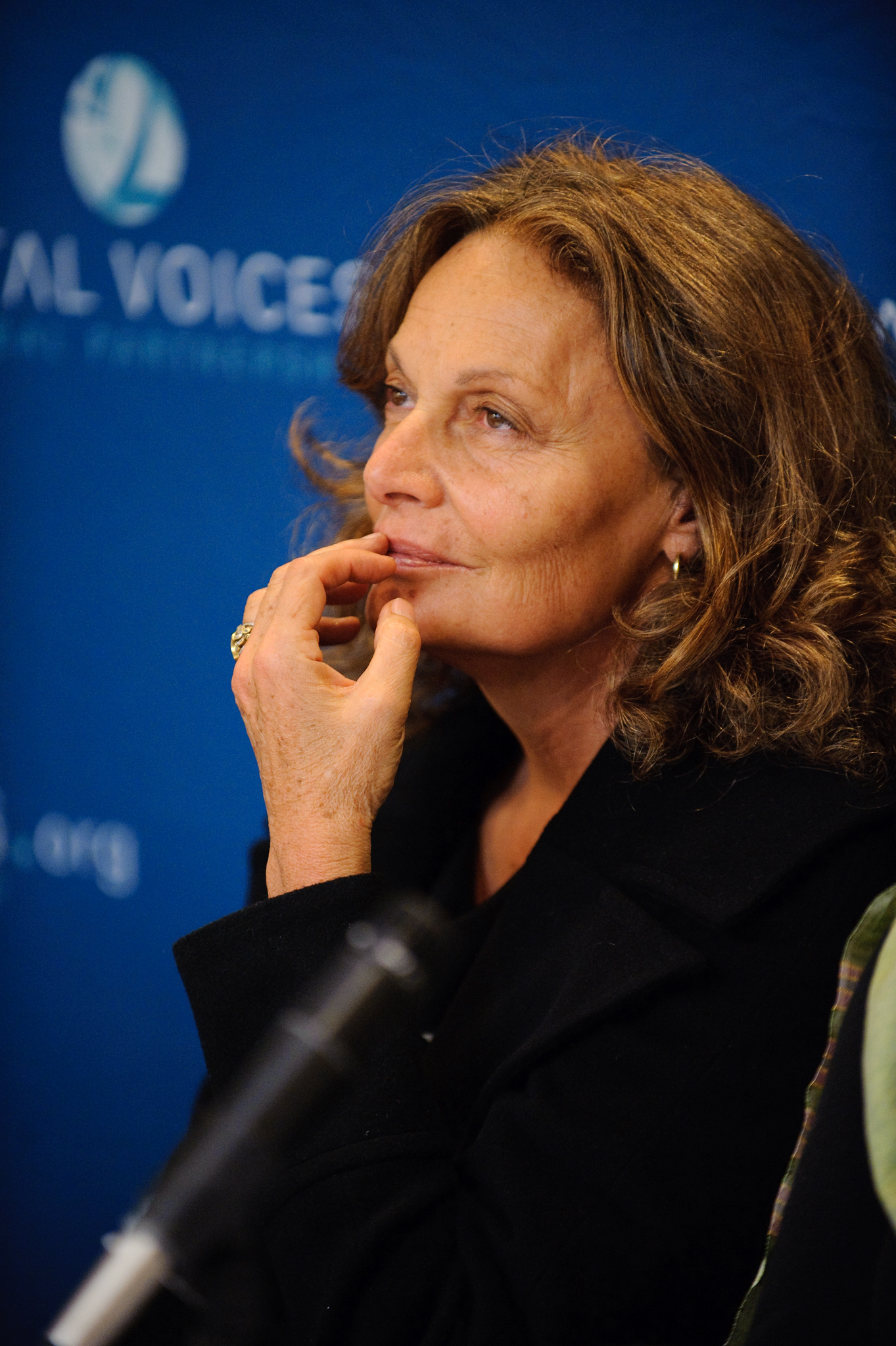 A woman with wavy brown hair sits in front of a blue backdrop, touching her chin thoughtfully and looking slightly upward.