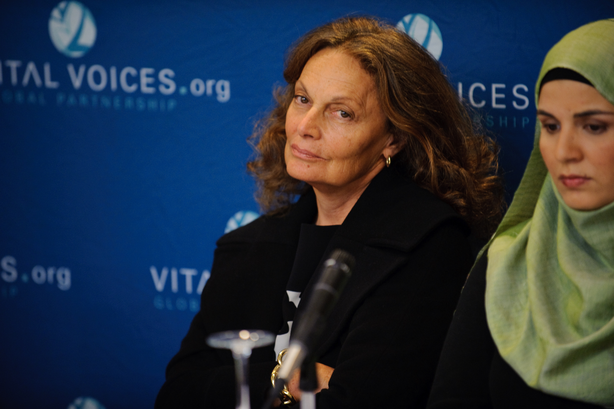 Two women sit in front of a blue Vital Voices.org backdrop; one wears a black coat, the other a green headscarf.