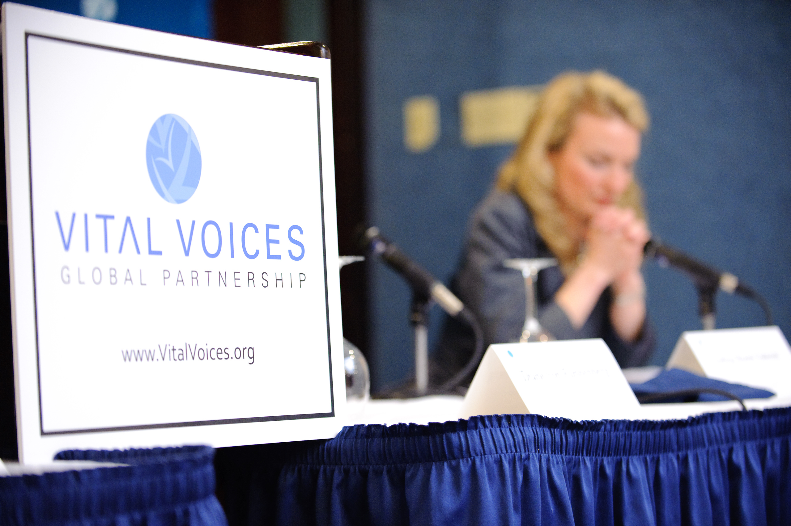 A Vital Voices Global Partnership sign is in focus on a table at a conference, with a woman sitting in the background, blurred, near microphones.
