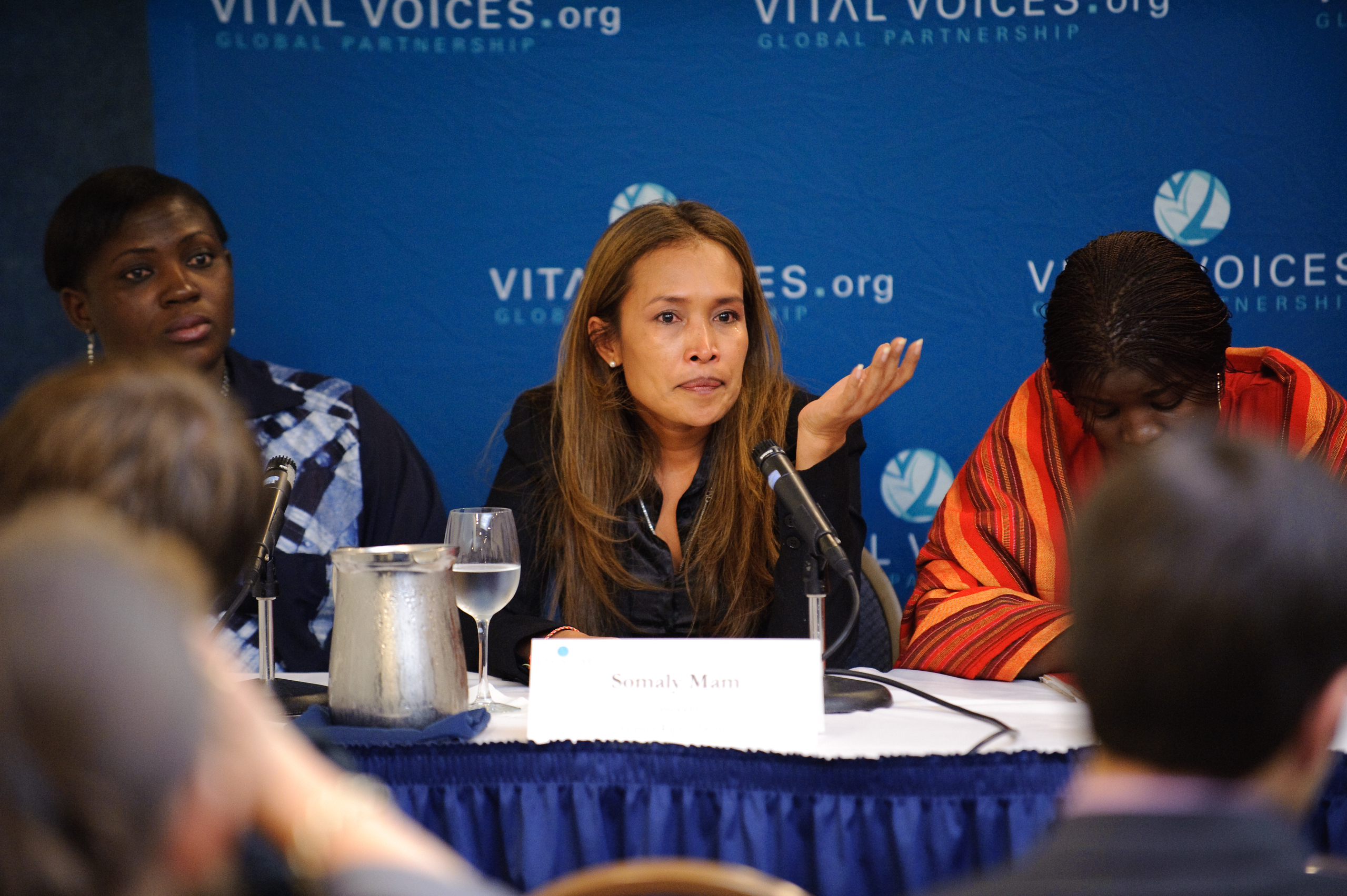 Three women sit at a panel table with microphones in front of a "Vital Voices" backdrop; the woman in the middle gestures while speaking to an audience.