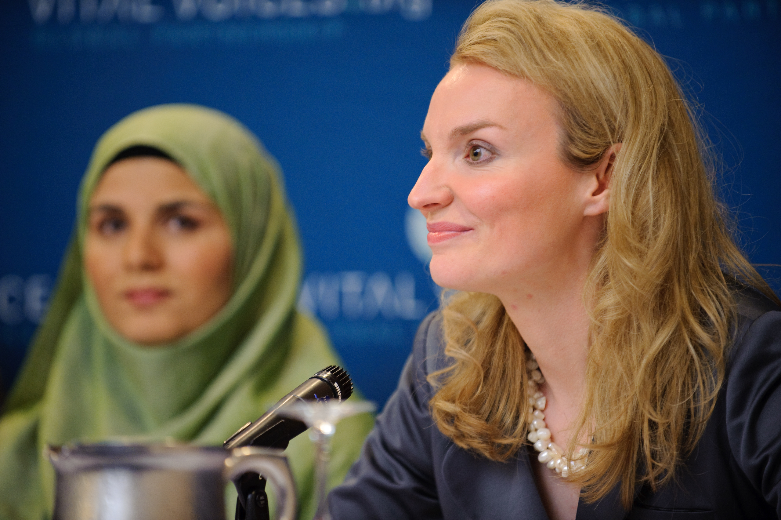 Two women sit at a table during a panel discussion, one wearing a green headscarf and the other in business attire, with a microphone and pitcher in front of them.