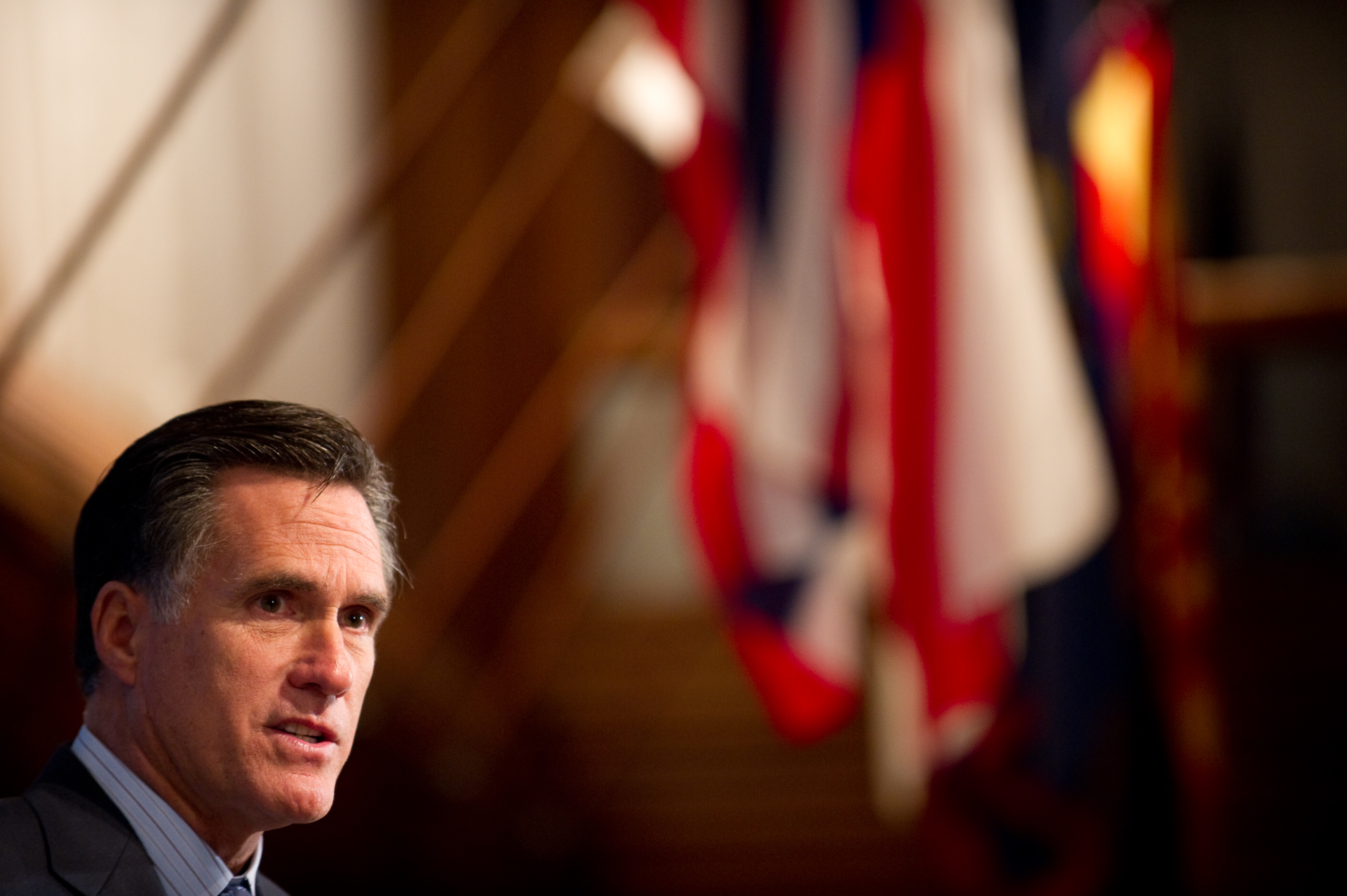 A man in a suit speaks in front of out-of-focus red, white, and blue flags in the background.