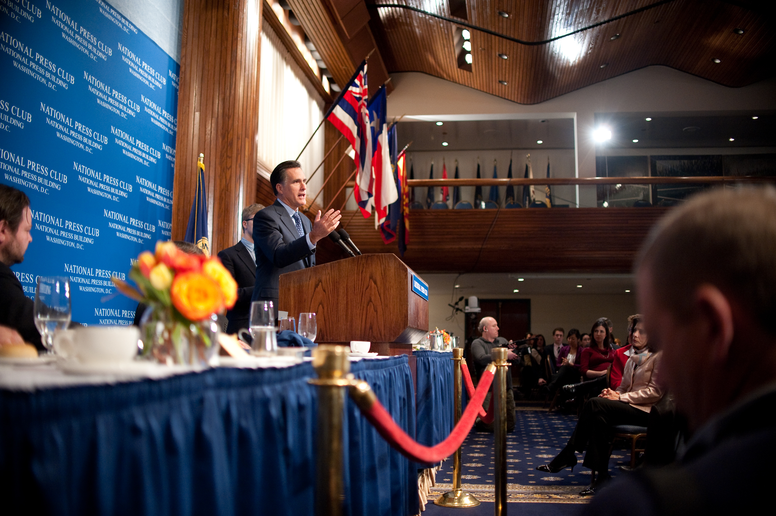 A man speaks at a podium on a stage with flags behind him at a formal event, facing an audience seated in a large room with wood-paneled ceiling and blue carpet.