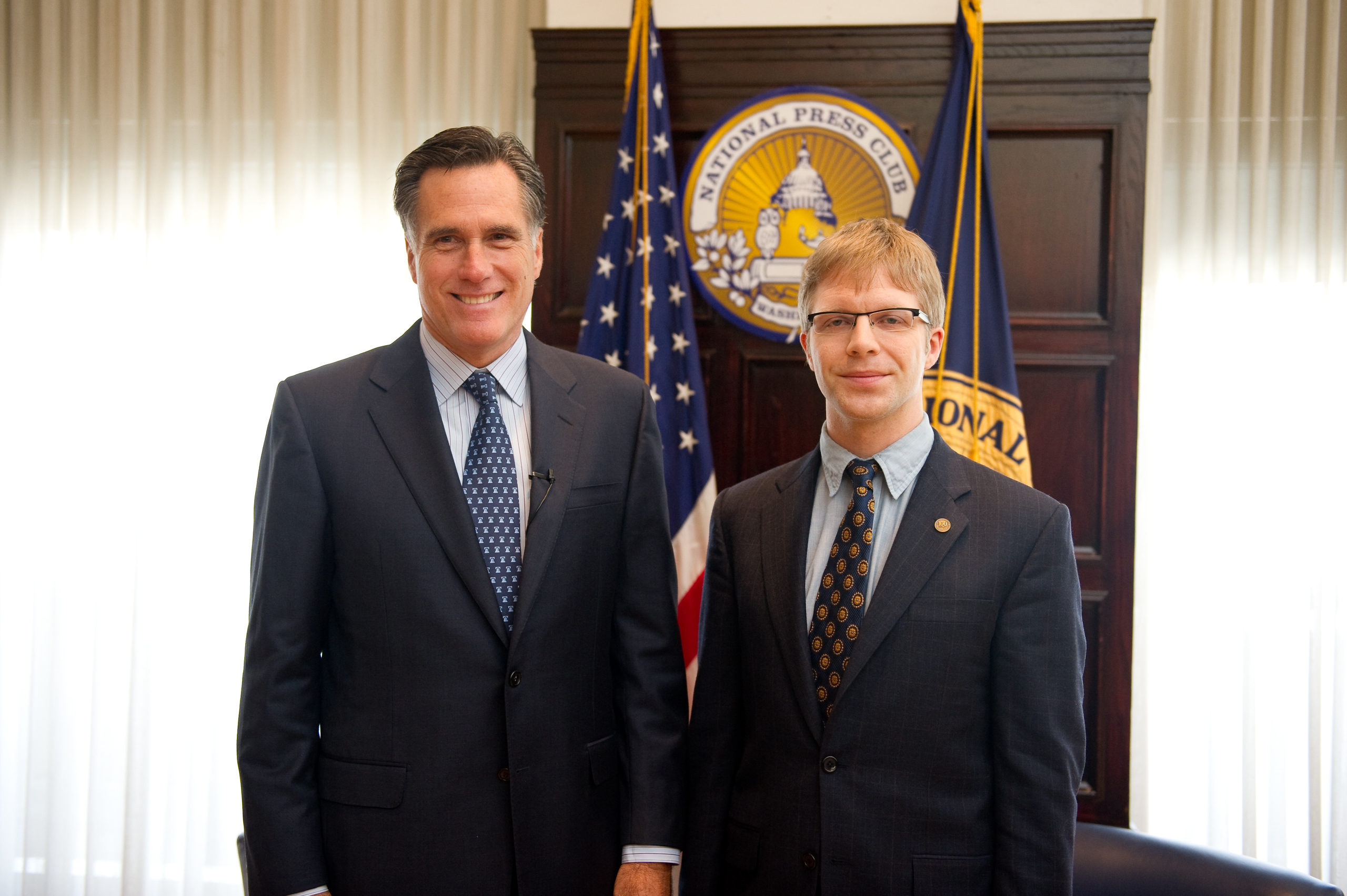 Two men in suits stand in front of the National Press Club emblem and an American flag in a formal indoor setting.