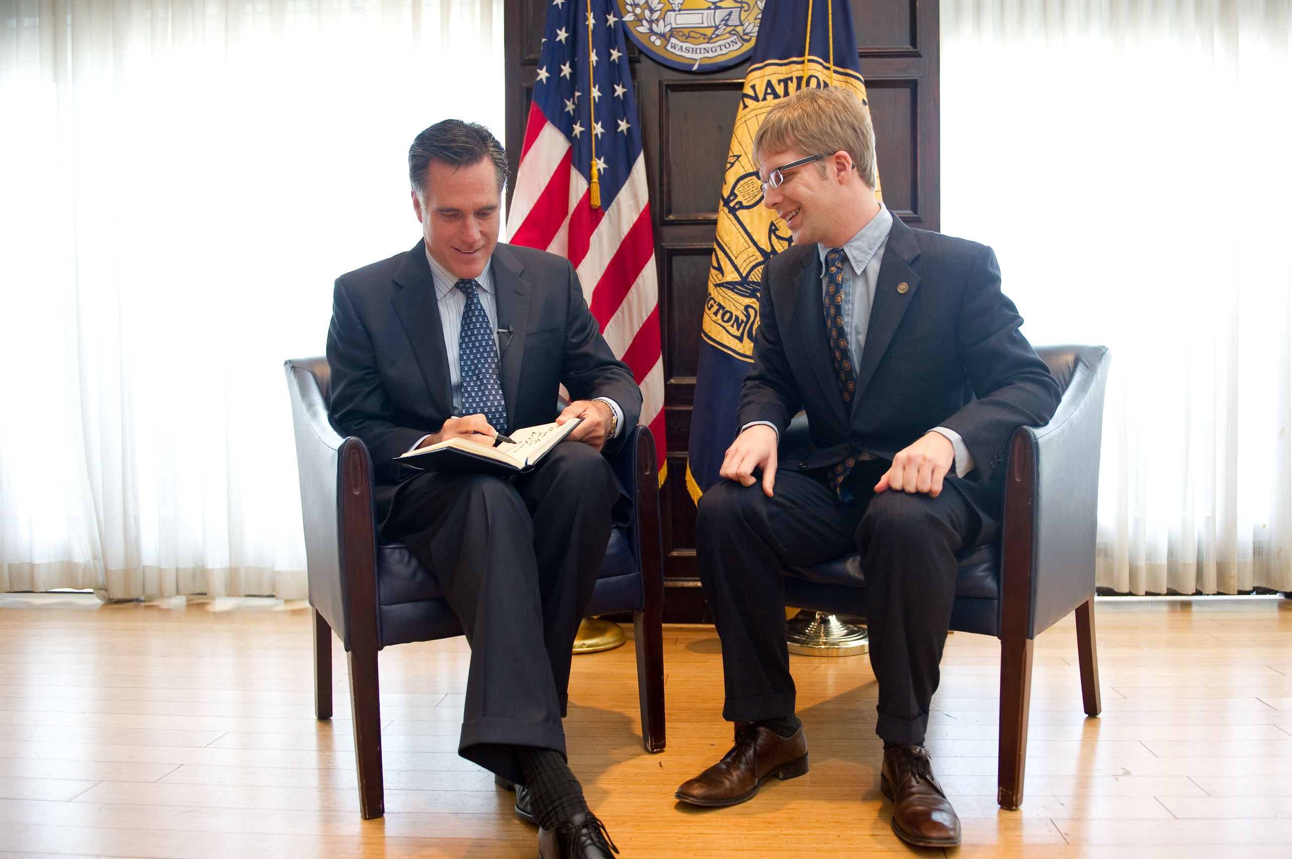 Two men in suits sit and talk in chairs, one signing a book. American flags and official emblems are visible in the background.