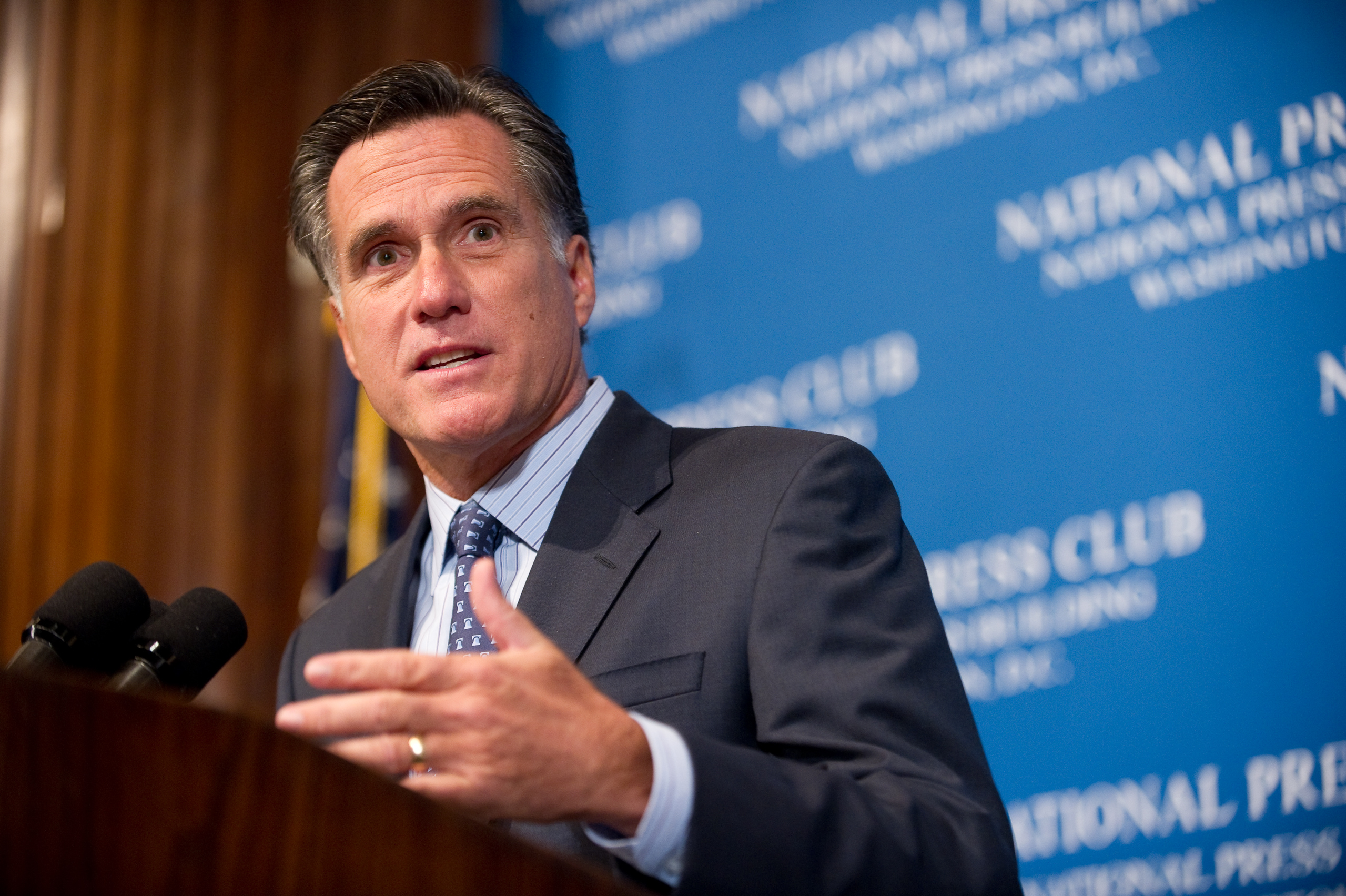 A man in a suit speaks at a podium with microphones in front of a blue National Press Club backdrop.